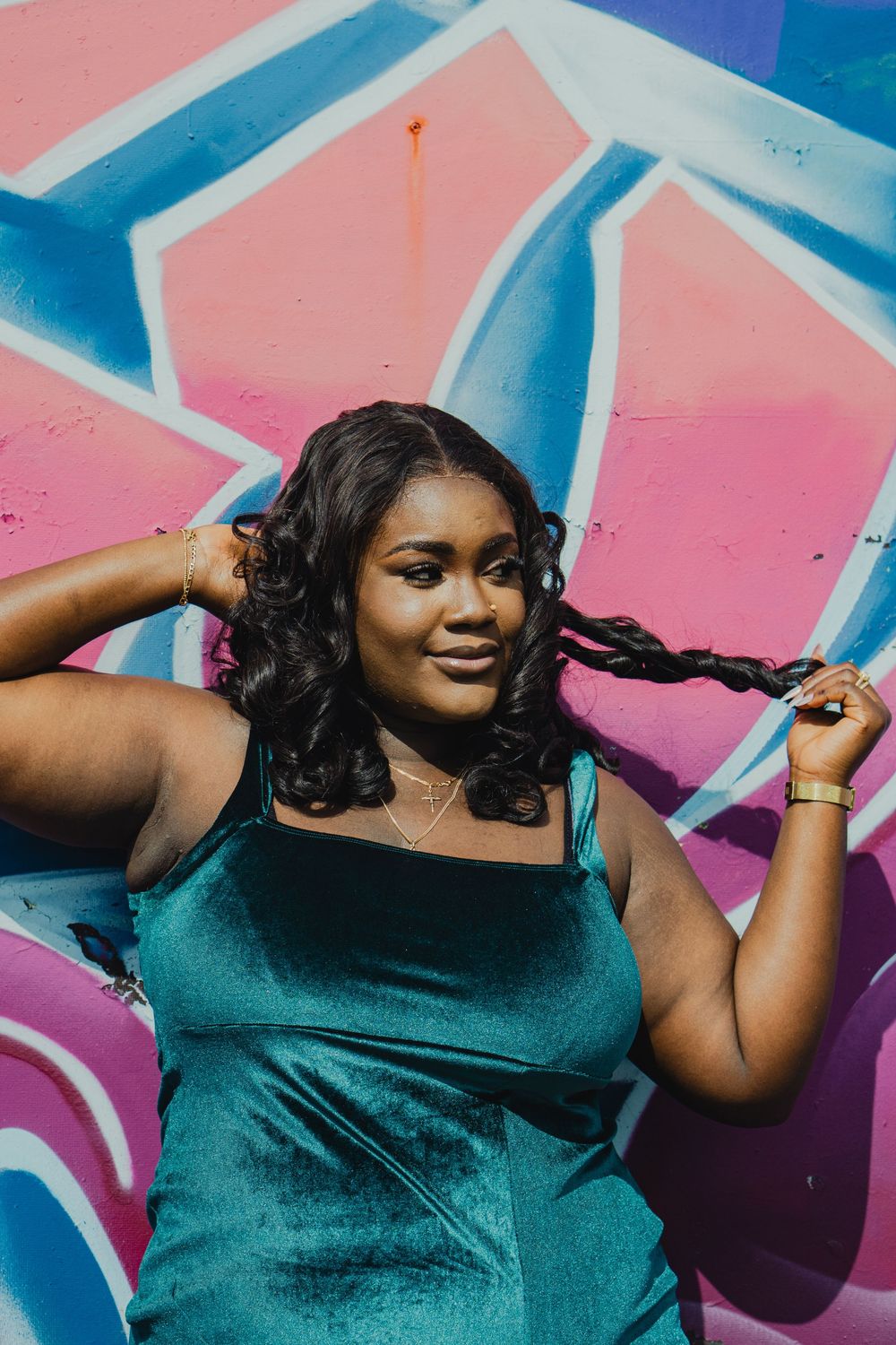 A young Black woman in a teal velvet dress poses confidently against a colorful pink and blue graffiti wall.