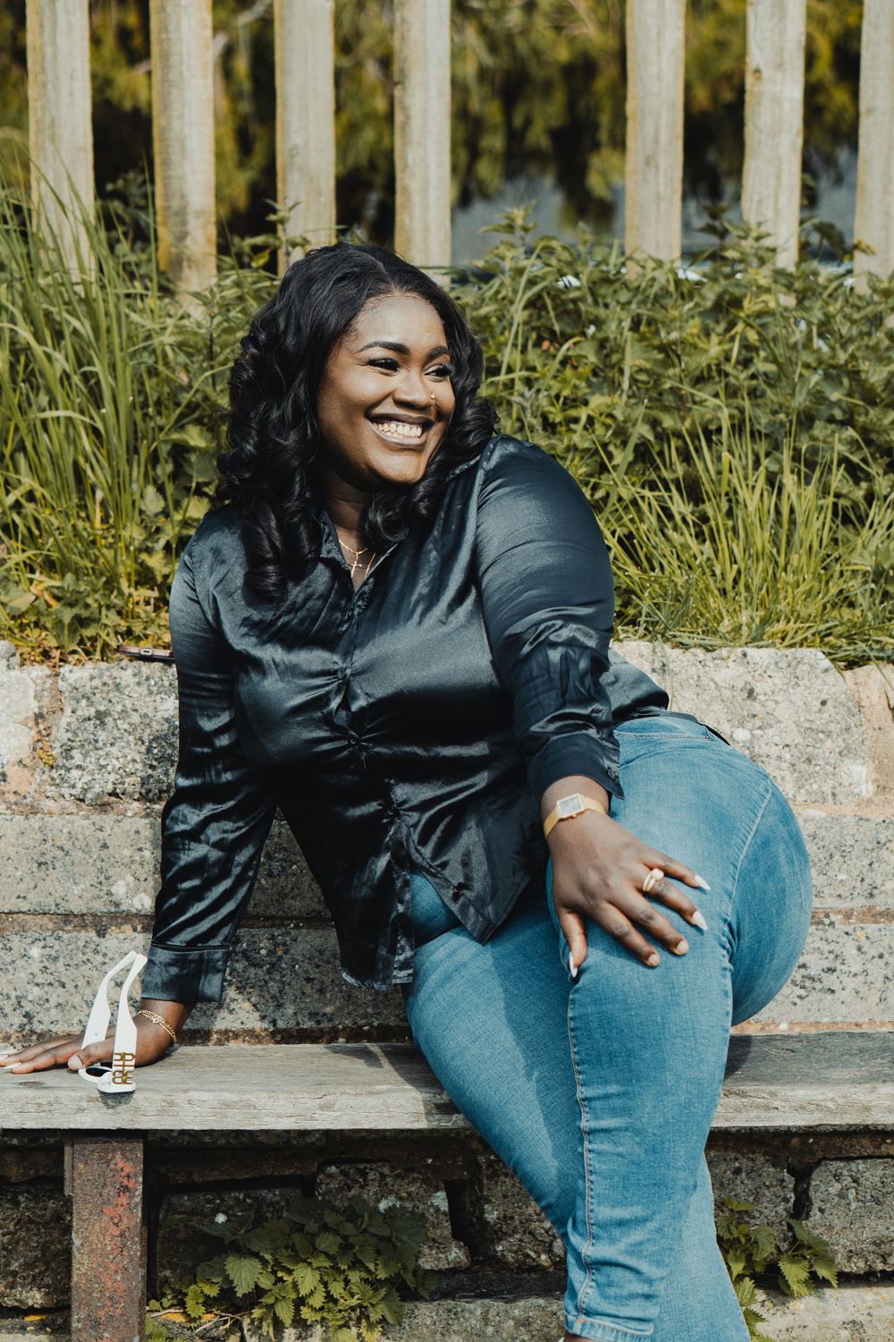 A smiling Black woman in a black satin shirt and jeans sits on a stone bench outdoors, posing confidently.