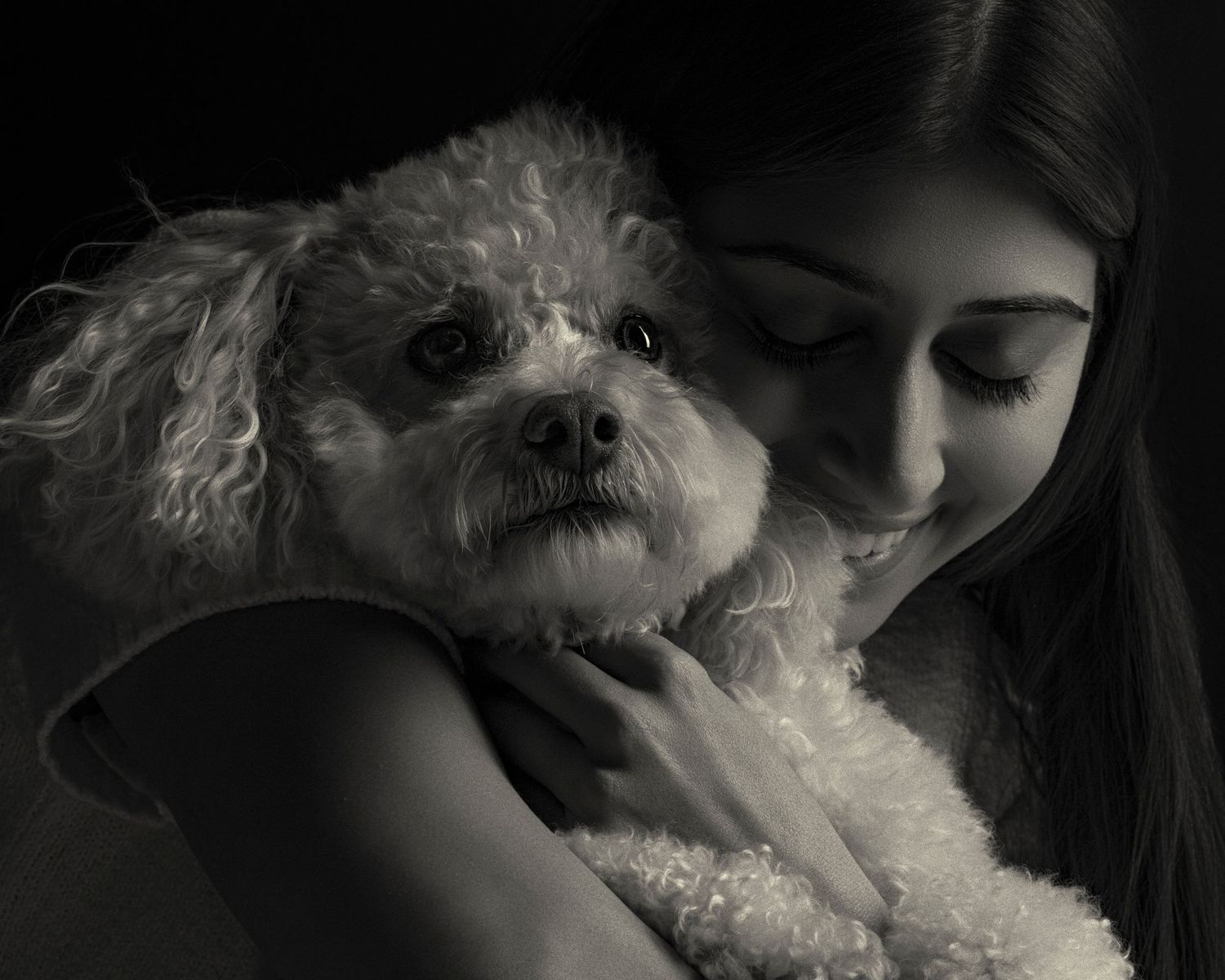 Black and white photo of a young girl tenderly embracing a fluffy white poodle dog in a dark studio setting.