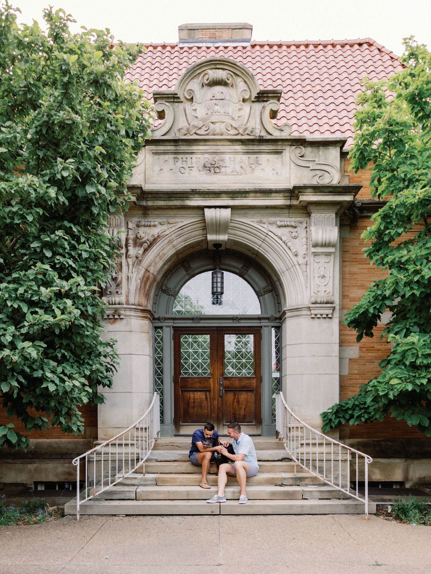 Couple sitting on steps of ornate stone building with arched wooden doors, lush green ivy, and red tile roof.