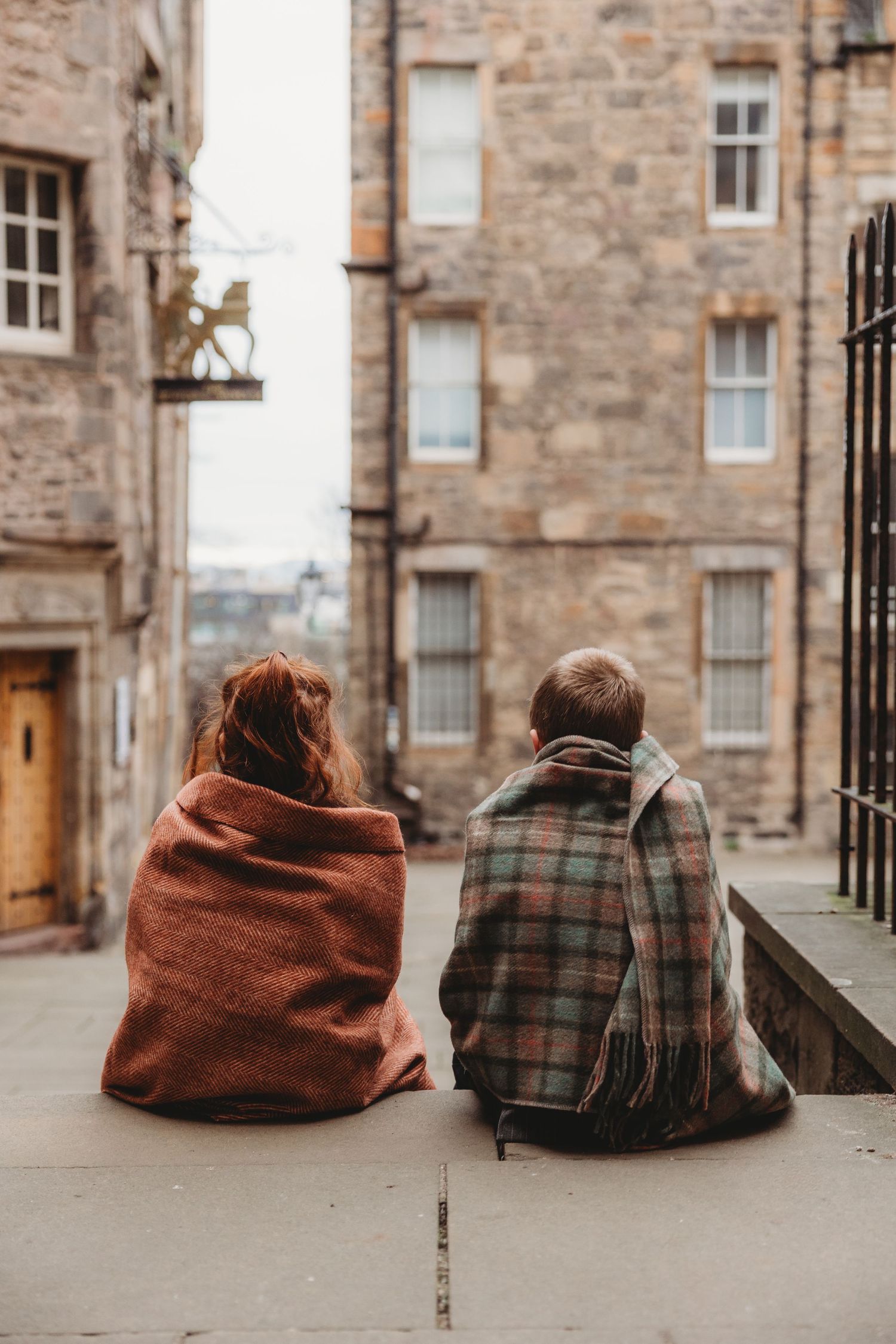 Two people wrapped in cozy blankets sit on stone steps in a historic cobblestone alley surrounded by old sandstone buildings.