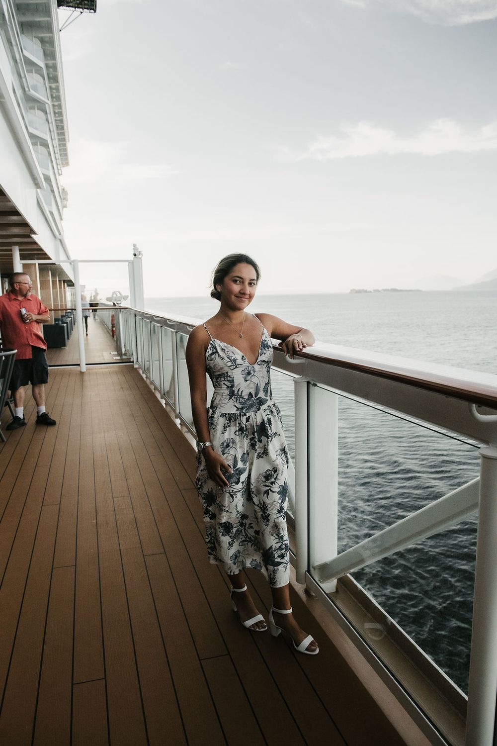 A smiling woman in a floral dress leans on a cruise ship railing with the ocean in the background.