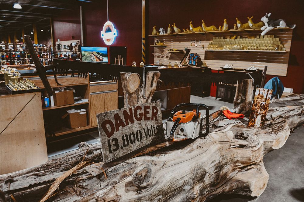 Interior of a shop displaying a large driftwood log with a Danger 13800 Volt warning sign and chainsaw on top.