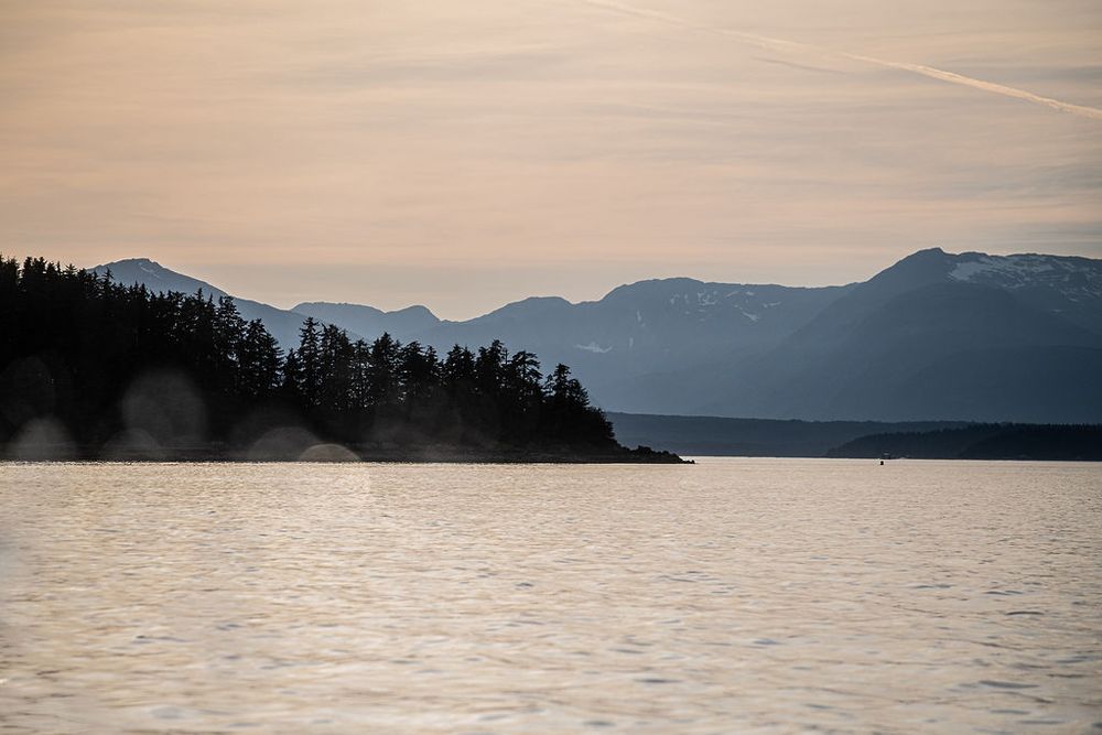 Silhouette of a forested island peninsula at dusk with layered mountain ridges in soft evening light over calm water.
