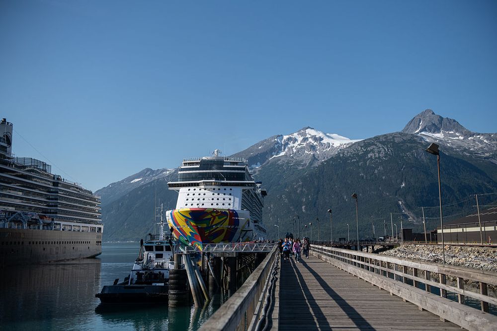 Norwegian cruise ship docked at Skagway, Alaska pier with dramatic snow-capped mountains under a clear blue sky.