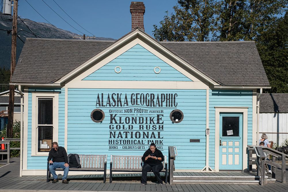 Two people sit on benches outside the light blue Alaska Geographic Klondike Gold Rush National Historical Park building.