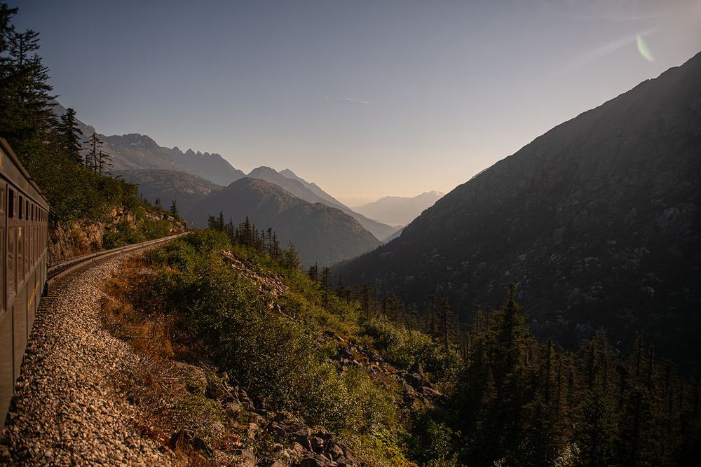 Historic narrow gauge railway winding along a steep mountain valley with evergreen forests and hazy peaks at golden hour.