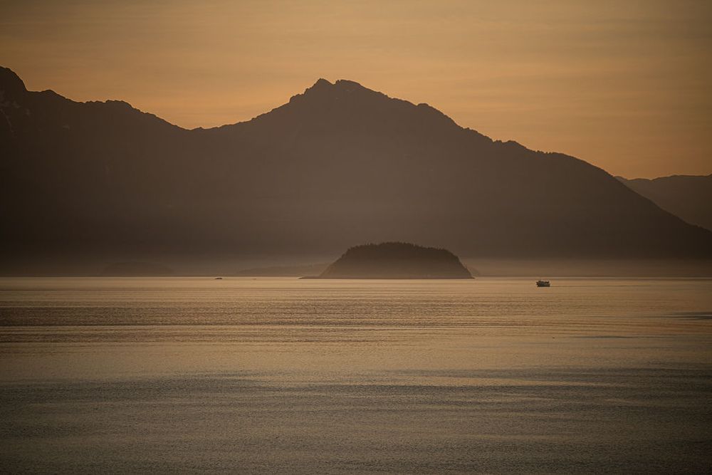 Layered mountain silhouettes rise above a misty golden bay at sunset with a small island.