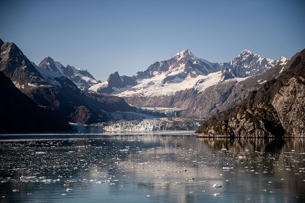 Snow-capped mountains and a glacier reflecting in the calm waters of an Alaskan fjord on a clear day.