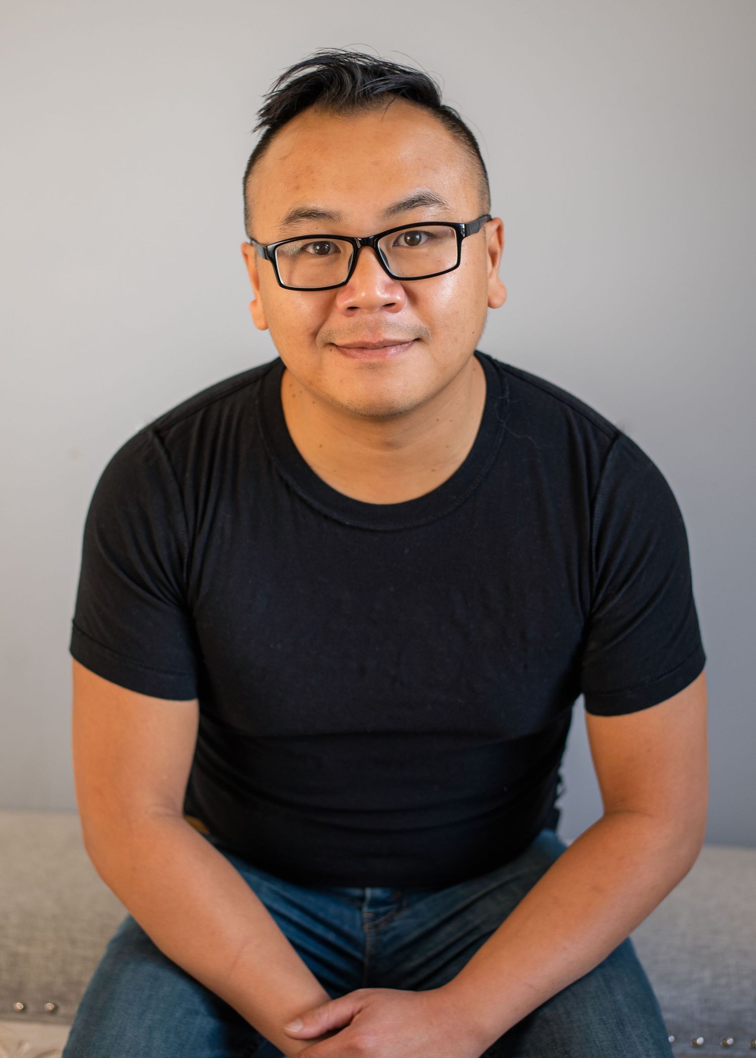 Man with glasses wearing a black t-shirt sitting casually, smiling in a modern indoor setting.