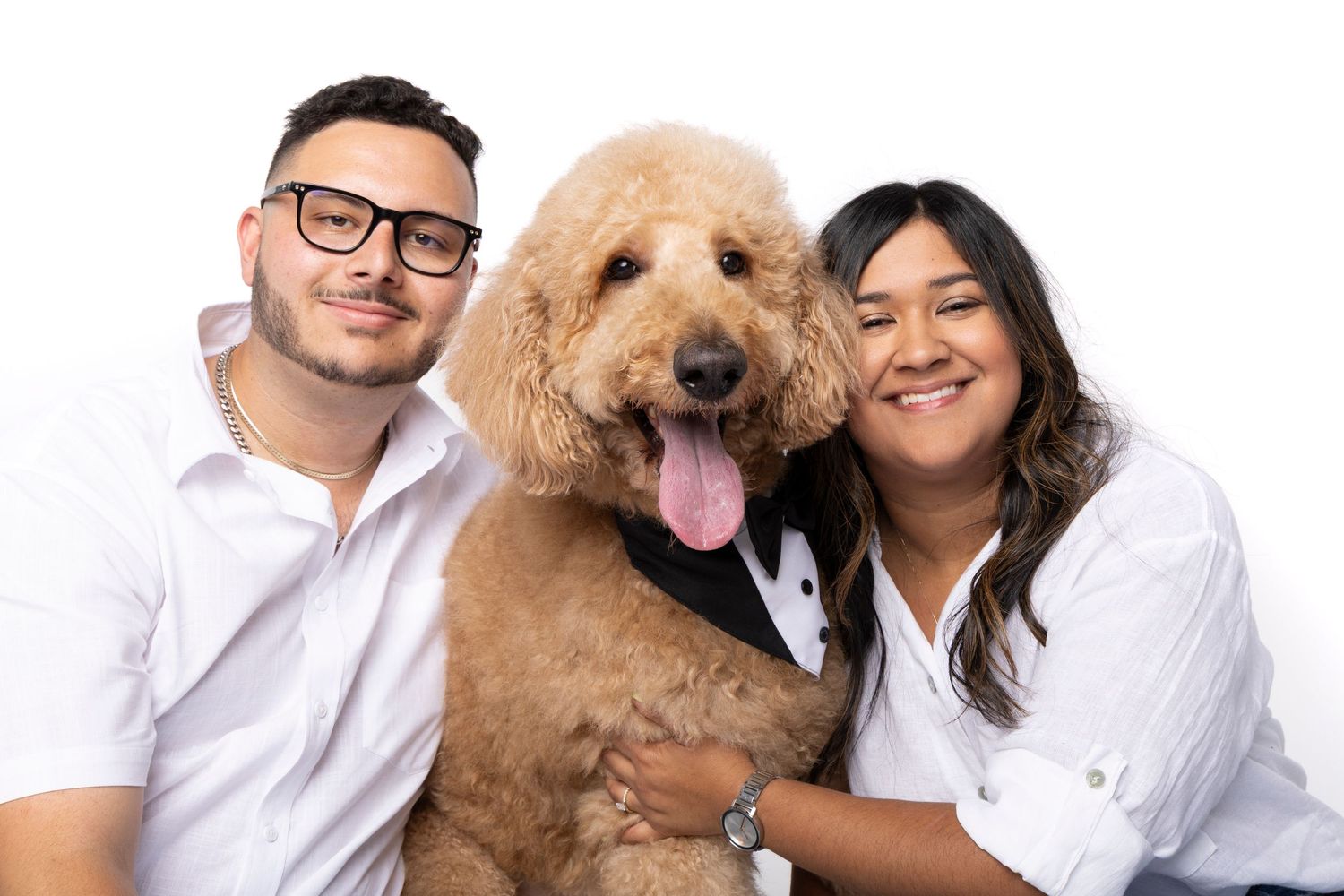 A couple in white shirts smile with their fluffy golden doodle dog wearing a tuxedo bib on a white background.