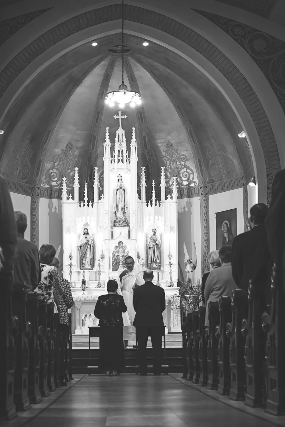 Black and white photo of a wedding ceremony at a Catholic church altar with ornate decor and guests standing in pews.
