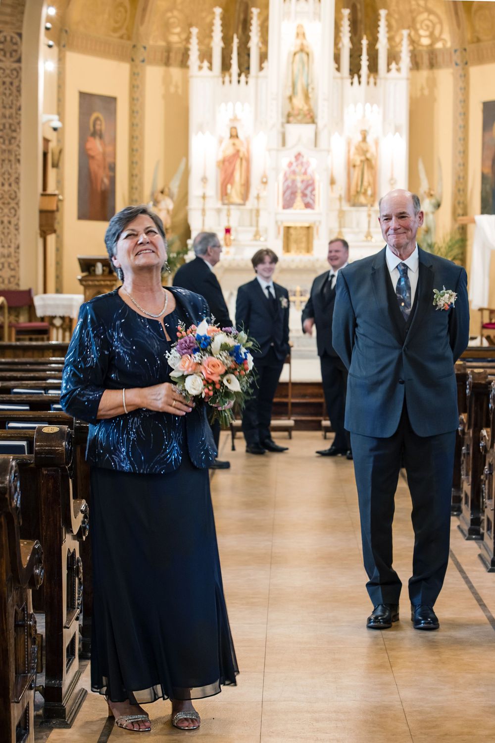 Couple walks down church aisle at wedding, woman in blue sequin dress holding bouquet, man in gray suit with boutonniere.