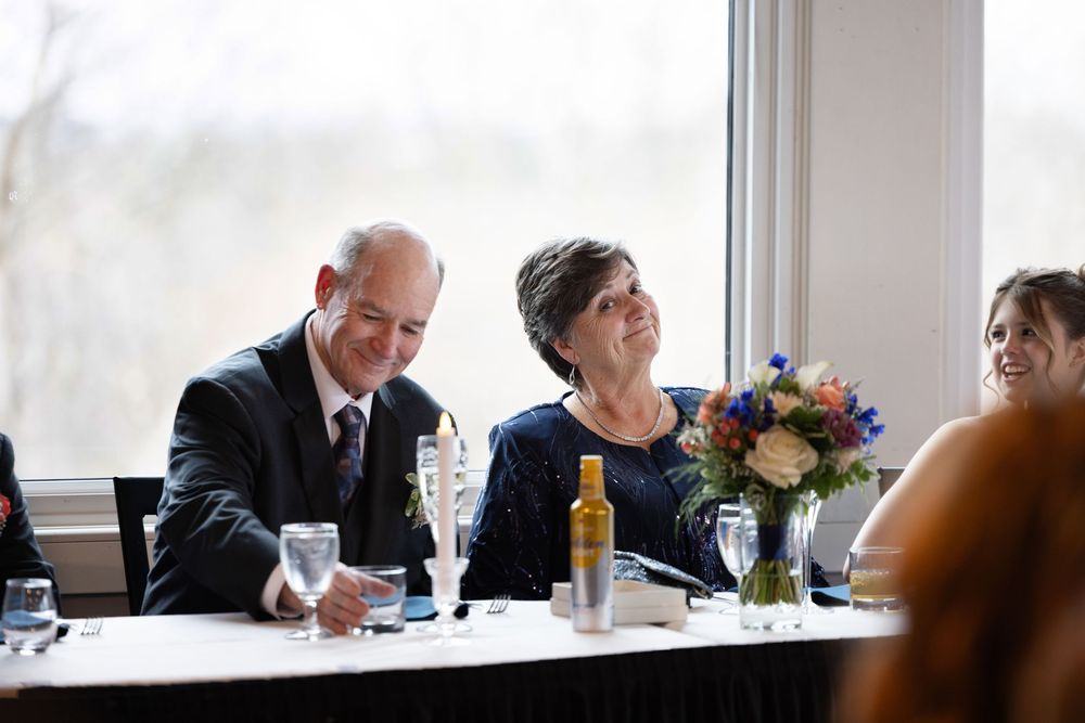 Couple smiling at wedding reception table with floral centerpiece and wine glasses near large windows.