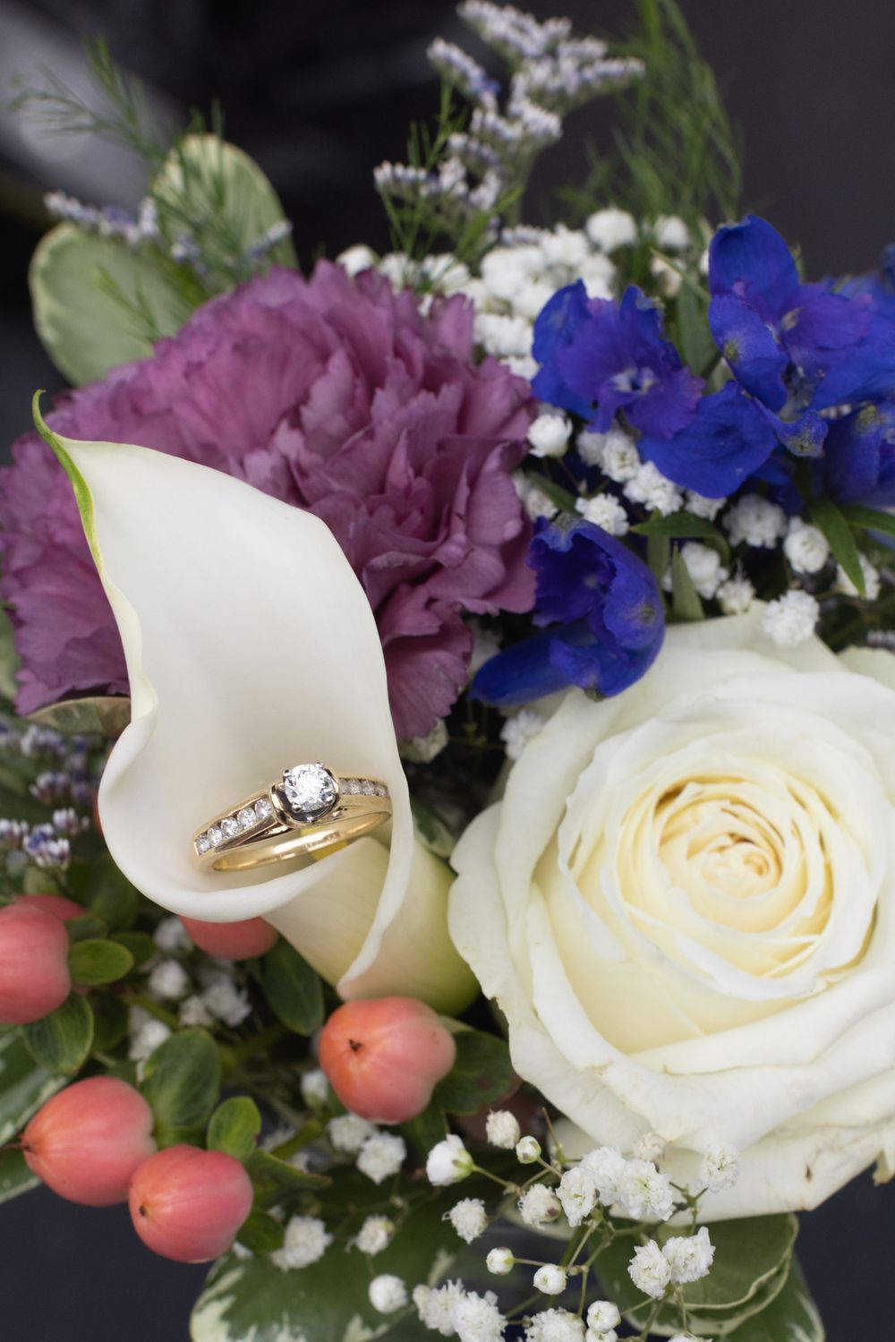 Close-up of gold diamond wedding rings resting on a bridal bouquet with white roses, calla lilies, and blue flowers.