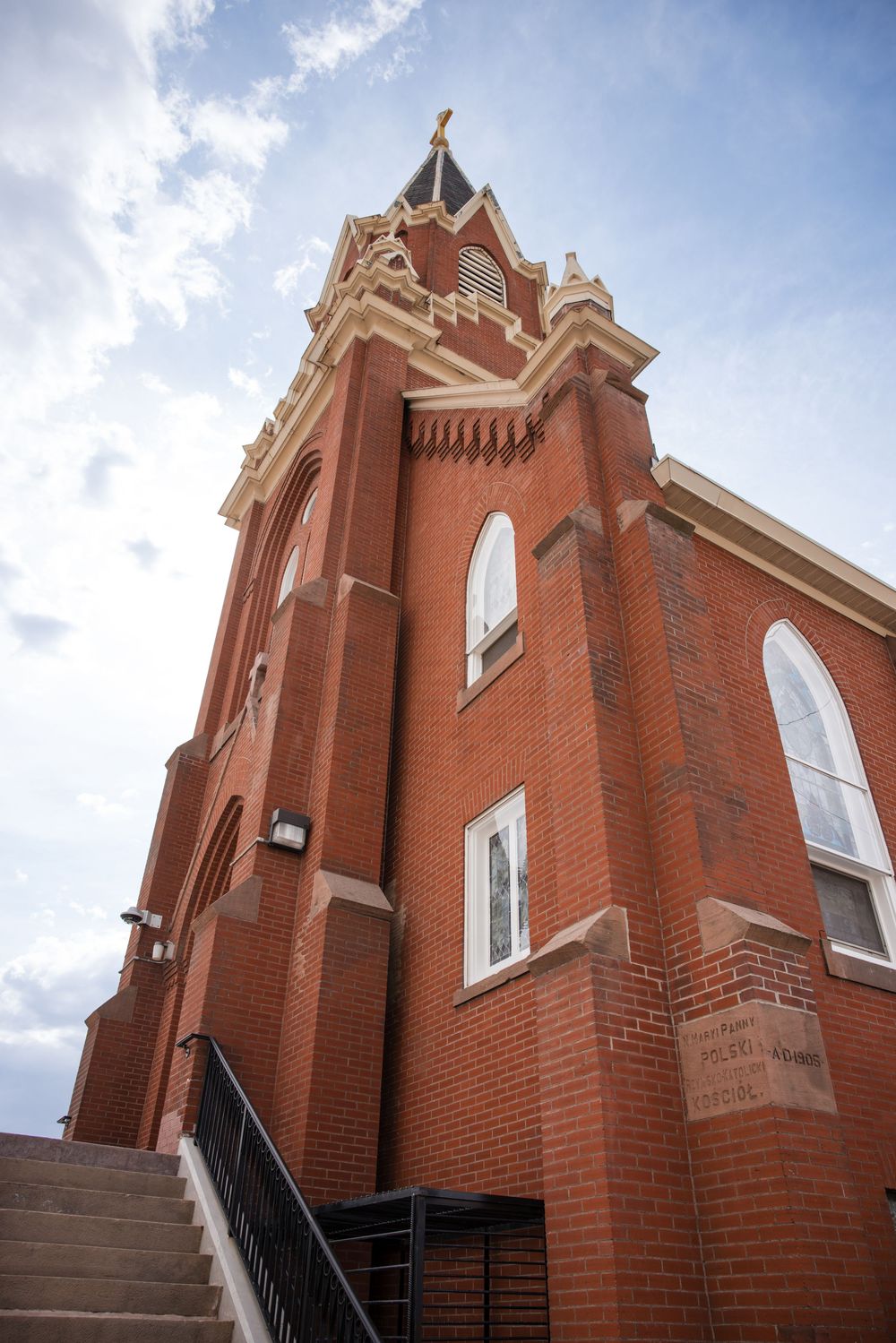 Historic red brick church with ornate Gothic tower, arched windows, and gold cross under a partly cloudy sky.