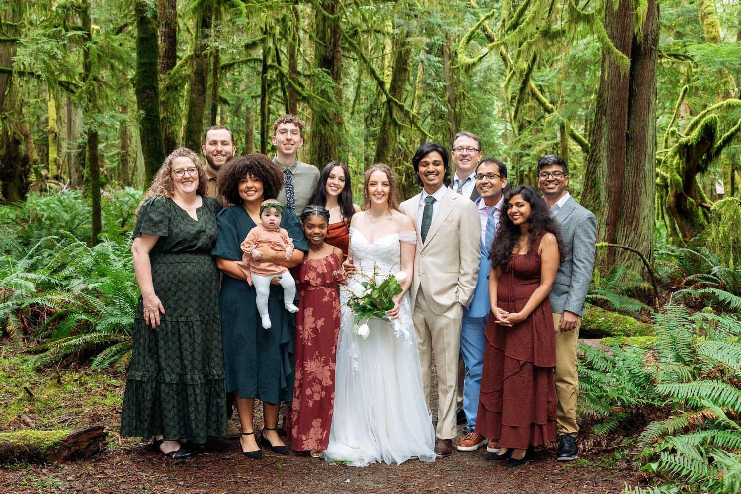 Wedding party posing together in a lush forest setting, bride in white gown with bouquet, groom in tan suit.