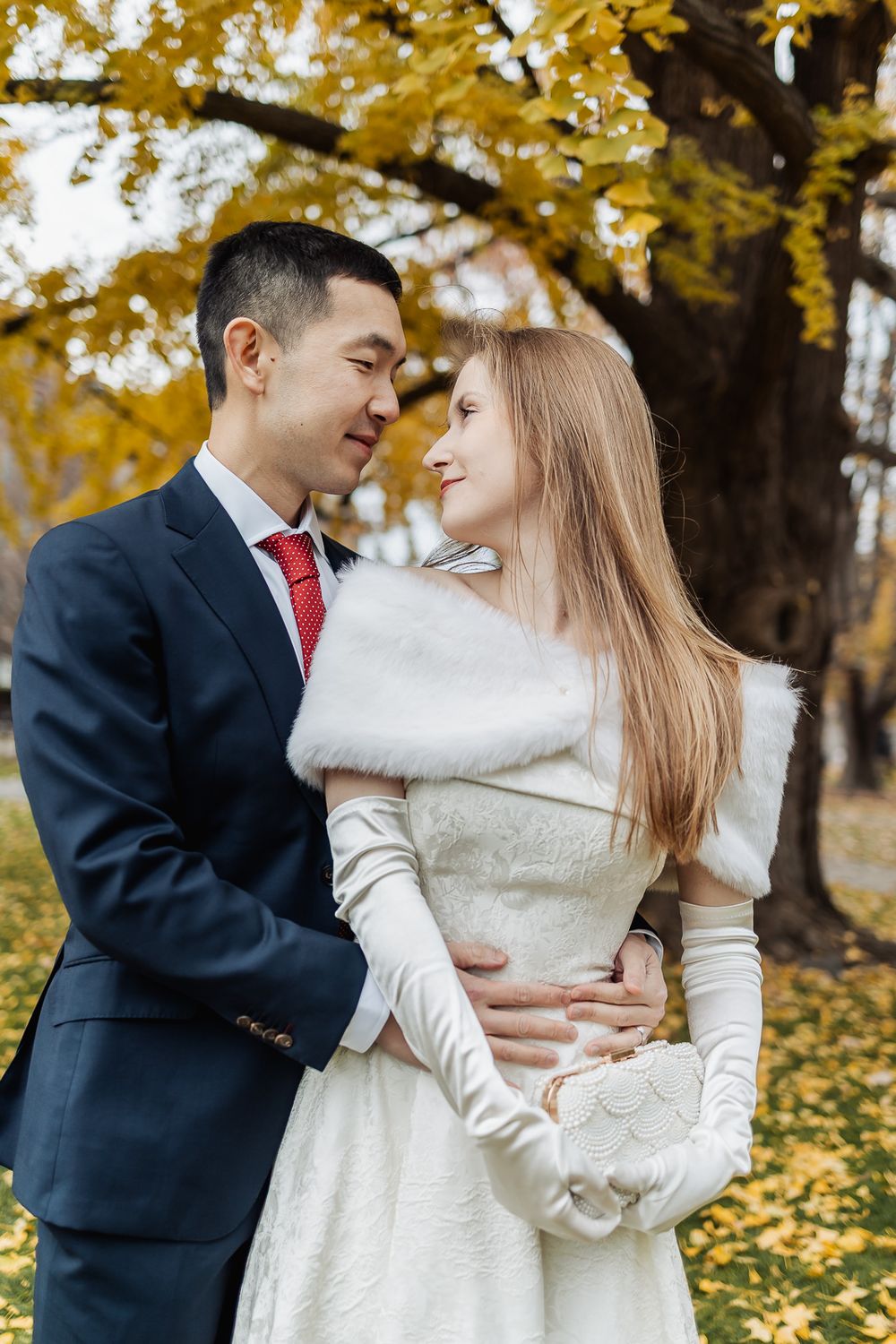 Newlywed couple embracing romantically under golden autumn trees, groom in navy suit with red tie, bride in white gown.