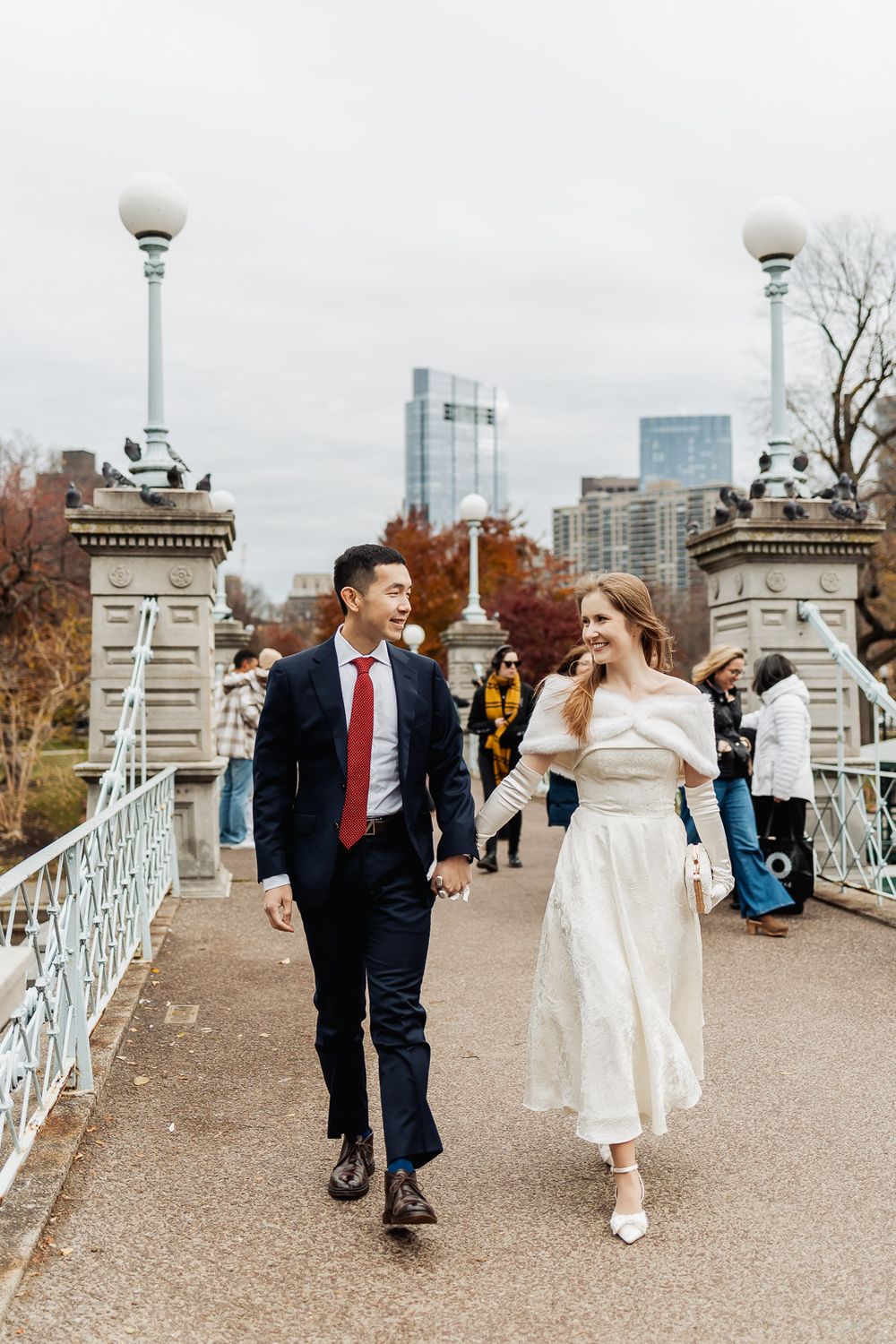 A couple in wedding attire holds hands walking on a bridge in Boston Public Garden with city skyline backdrop.