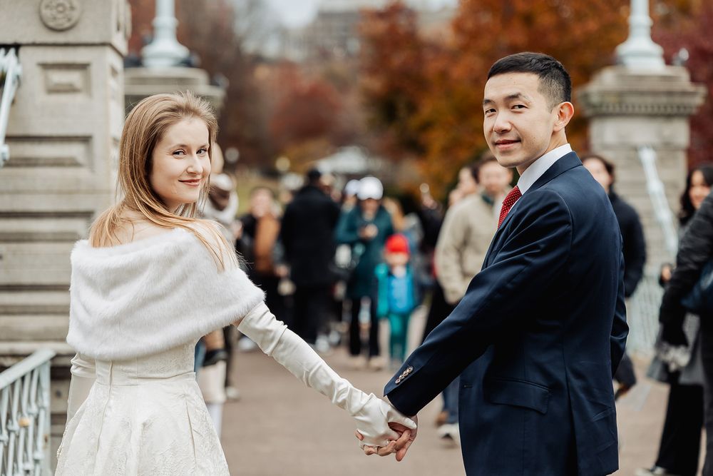 Couple in wedding attire holding hands outdoors in autumn park, bride in white wrap smiling over shoulder.