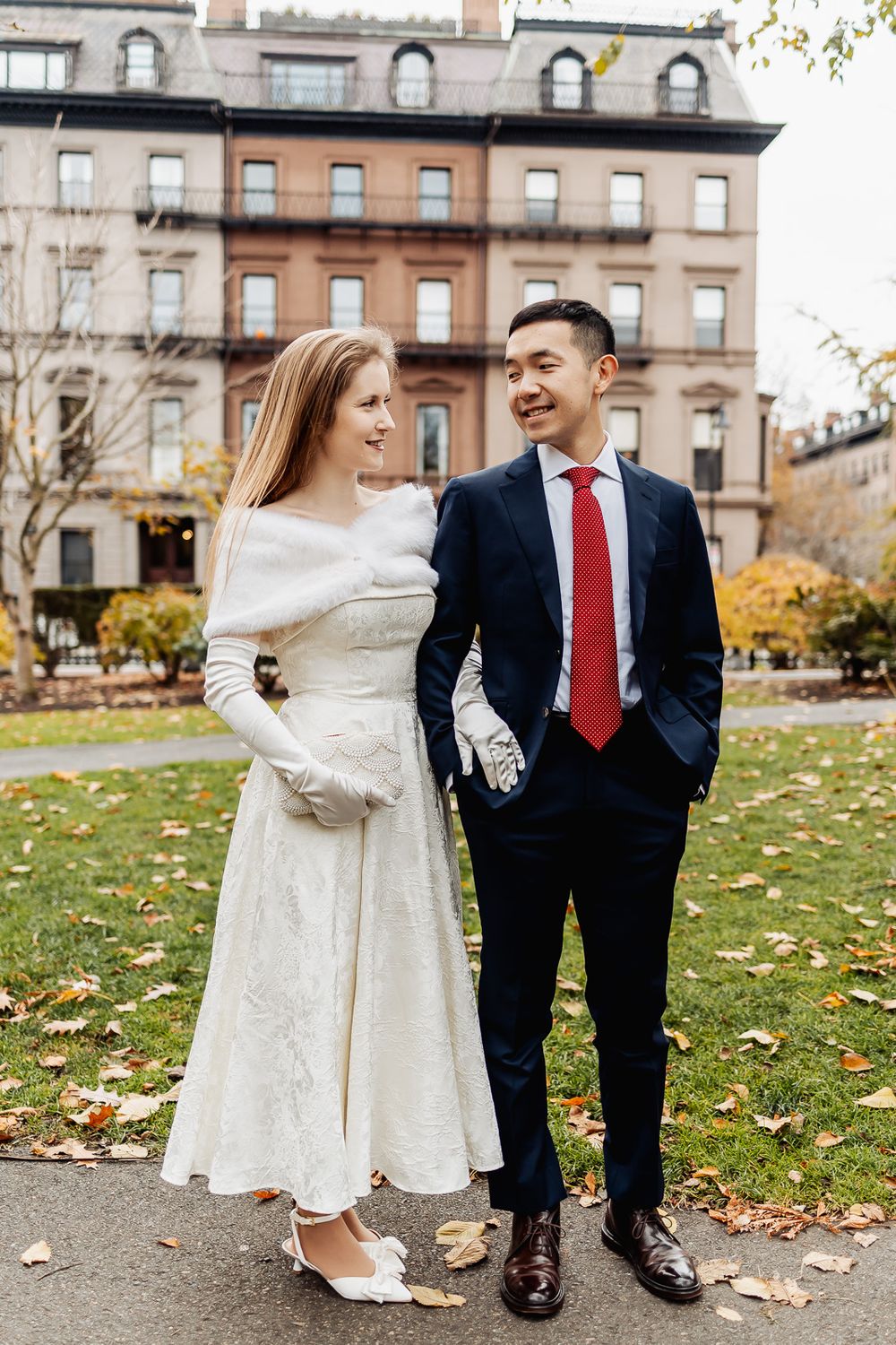 A couple in wedding attire smiles at each other outdoors in autumn, she in white dress, he in navy suit with red tie.