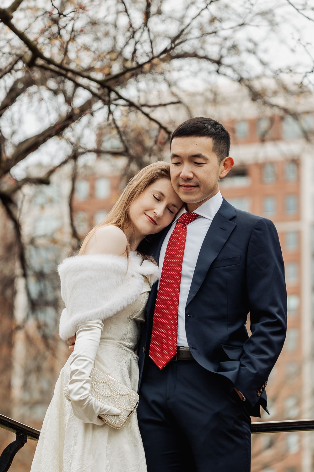 Elegant couple in winter wedding attire embracing outdoors, bride in white dress with fur wrap, groom in navy suit with red tie.