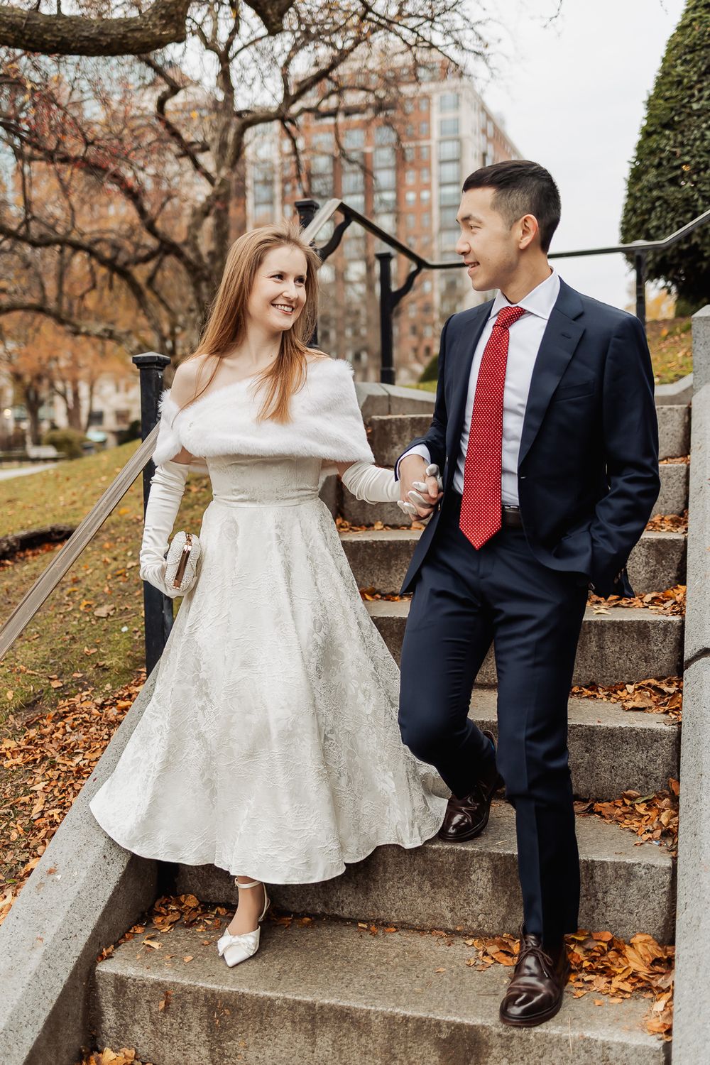 Bride and groom holding hands on stone steps in a park during autumn, wearing white dress and navy suit with red tie.