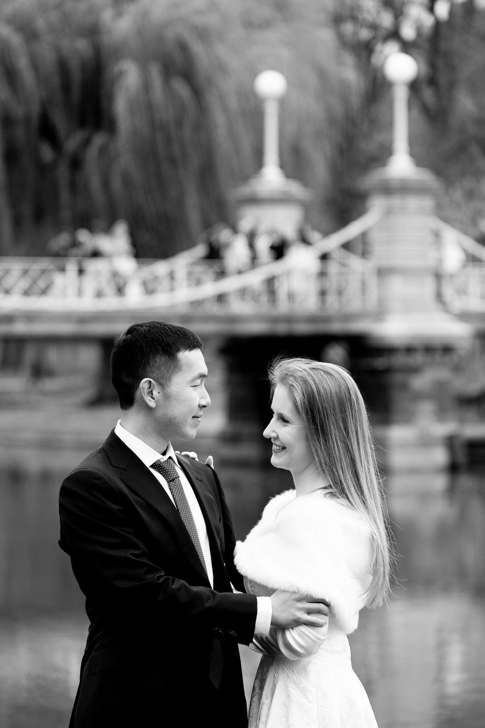Black and white photo of wedding couple embracing near bridge and pond, bride in white fur wrap.