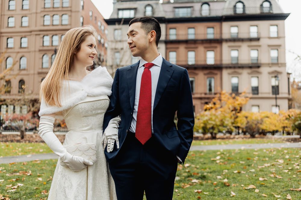 A couple in wedding attire share a romantic moment outdoors, with the bride in white gloves and groom in a navy suit with red tie.