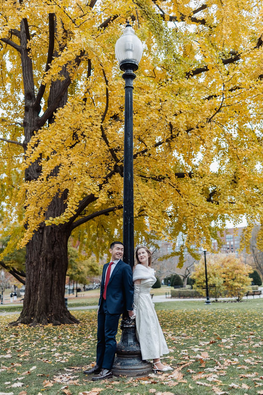 Couple poses by a black lamppost under a vibrant yellow ginkgo tree in a park during autumn.