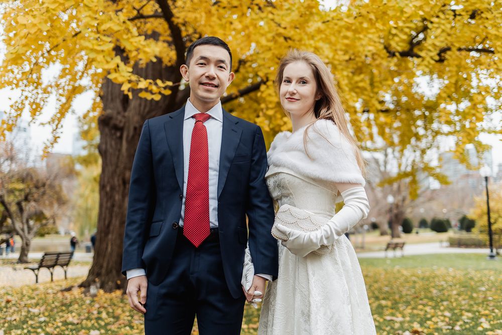 Couple in wedding attire holding hands in autumn park, groom in navy suit red tie, bride in white dress.