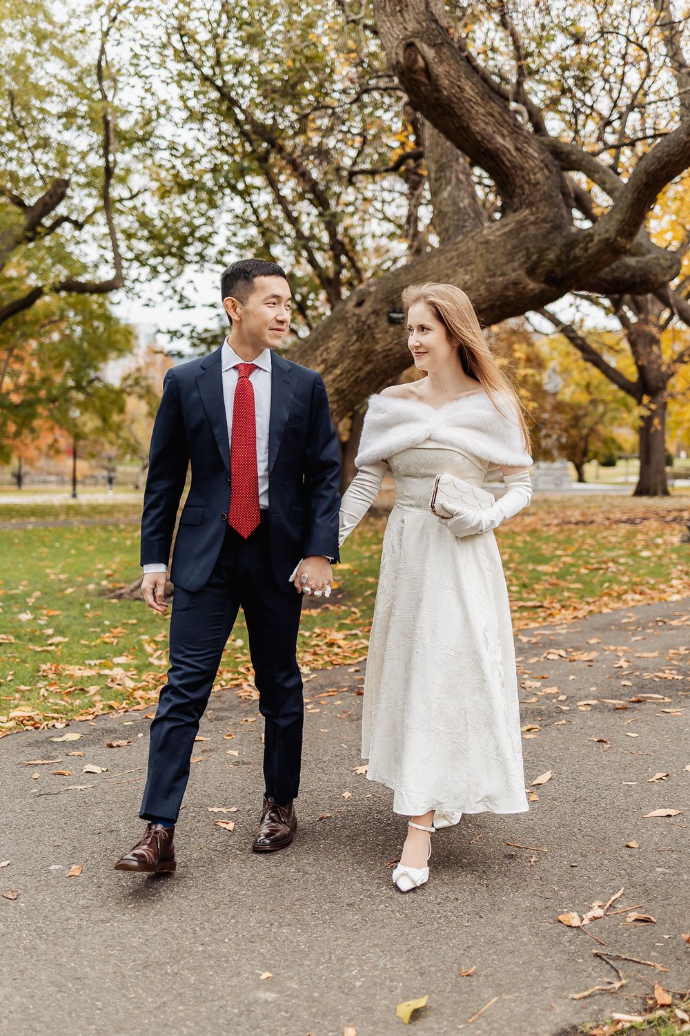 Couple in wedding attire walking hand in hand through an autumn park, groom in navy suit, bride in white dress.