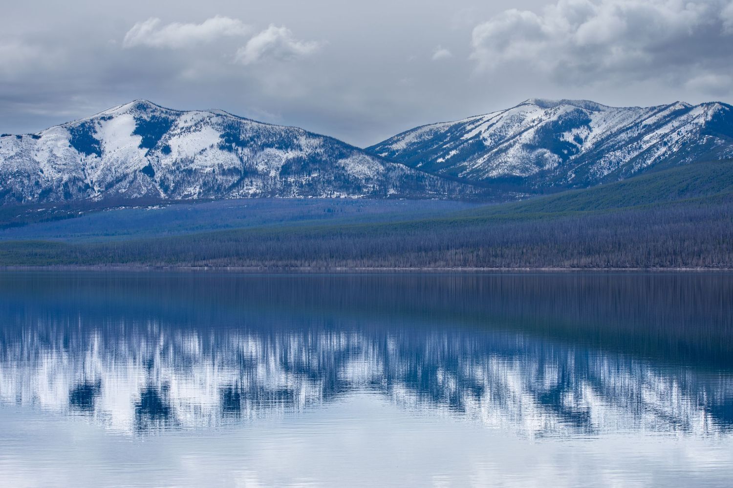 Snow-capped mountains reflect perfectly in the calm blue waters of a serene alpine lake under cloudy skies.