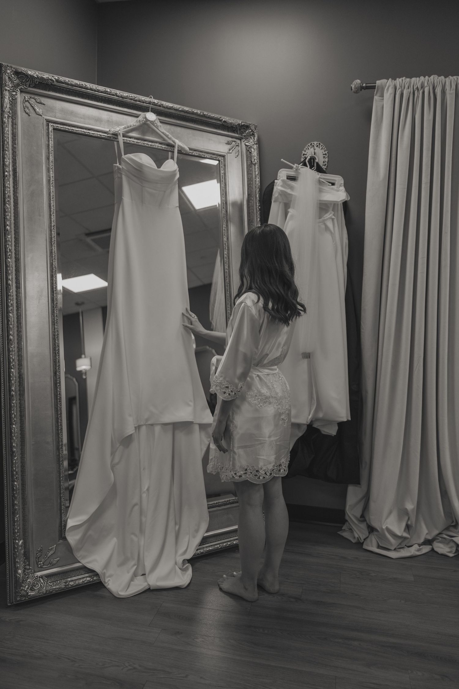 Black and white photograph of wedding dresses hanging in a bridal shop fitting room with ornate mirrors.