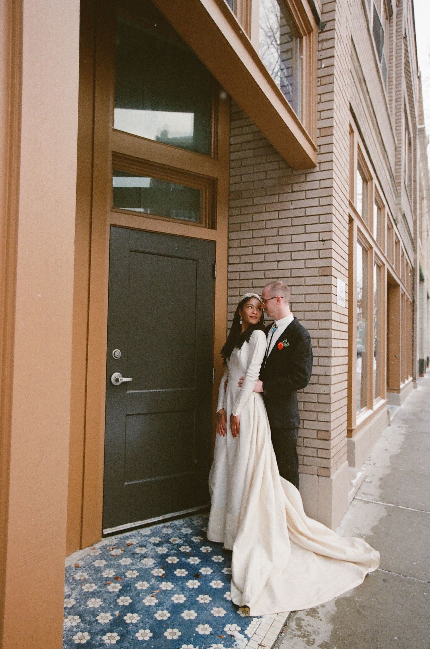 A bride and groom pose elegantly by a dark door on a brick building, her long white gown trailing on blue tile.