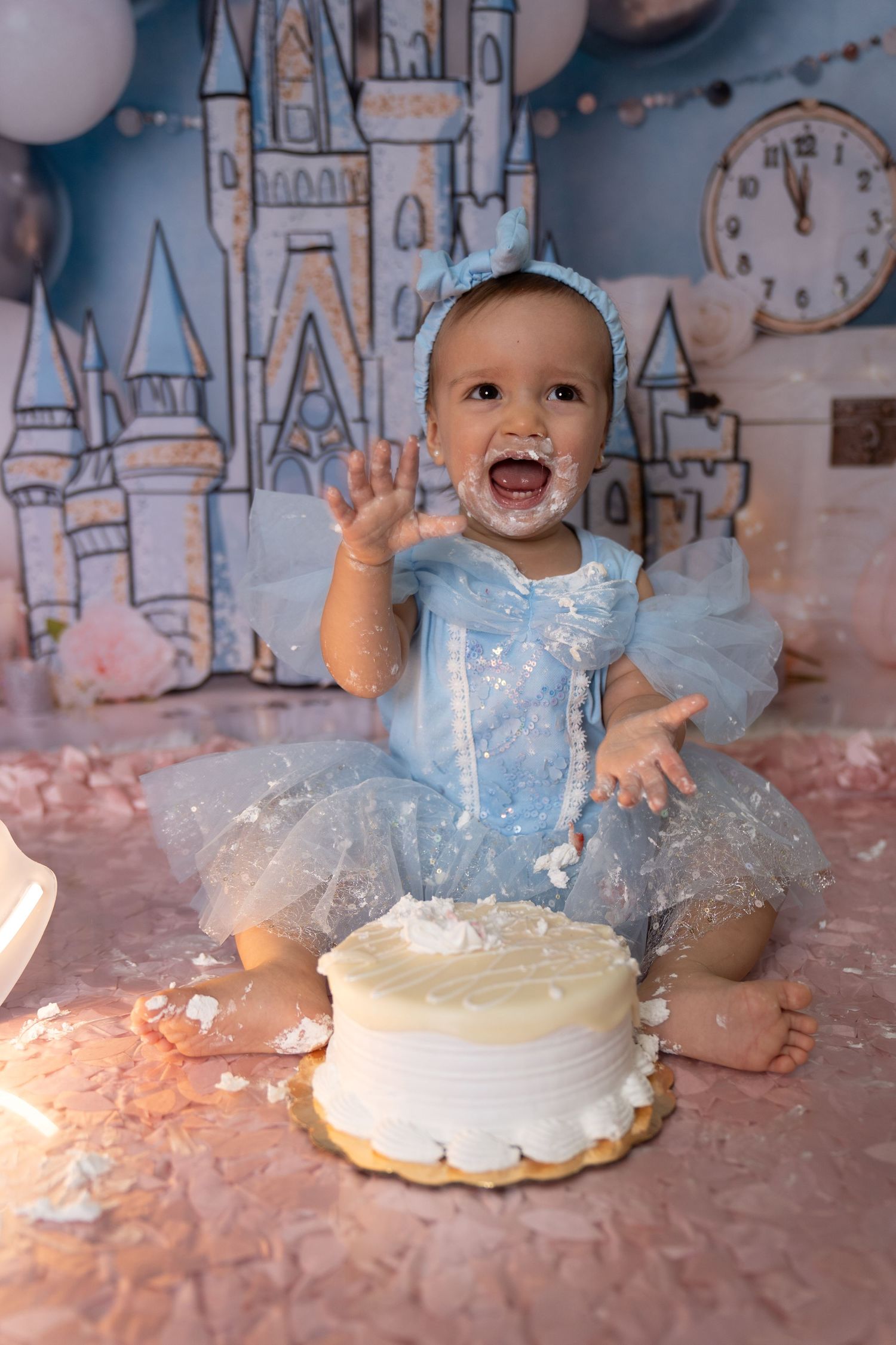 Baby girl in blue tutu dress celebrates first birthday cake smash with castle backdrop and balloon decorations.