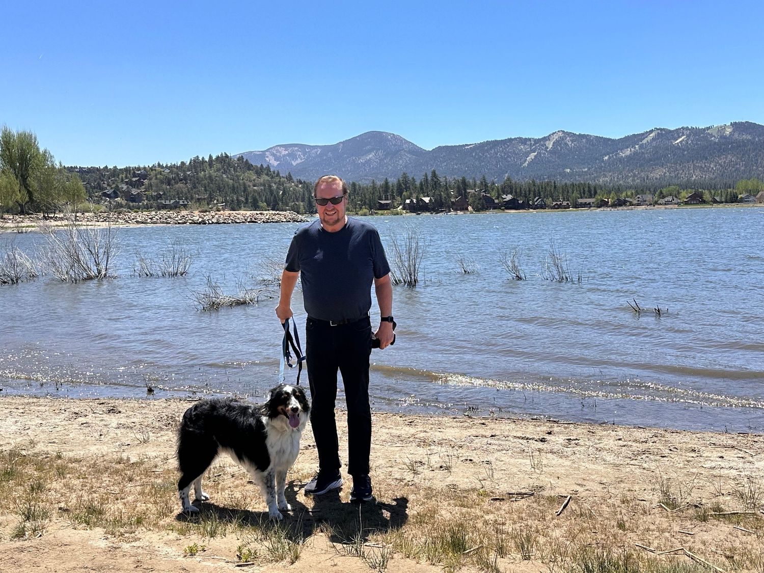 Man and border collie dog standing on sandy beach shore with mountain lake and snow-capped peaks in background.