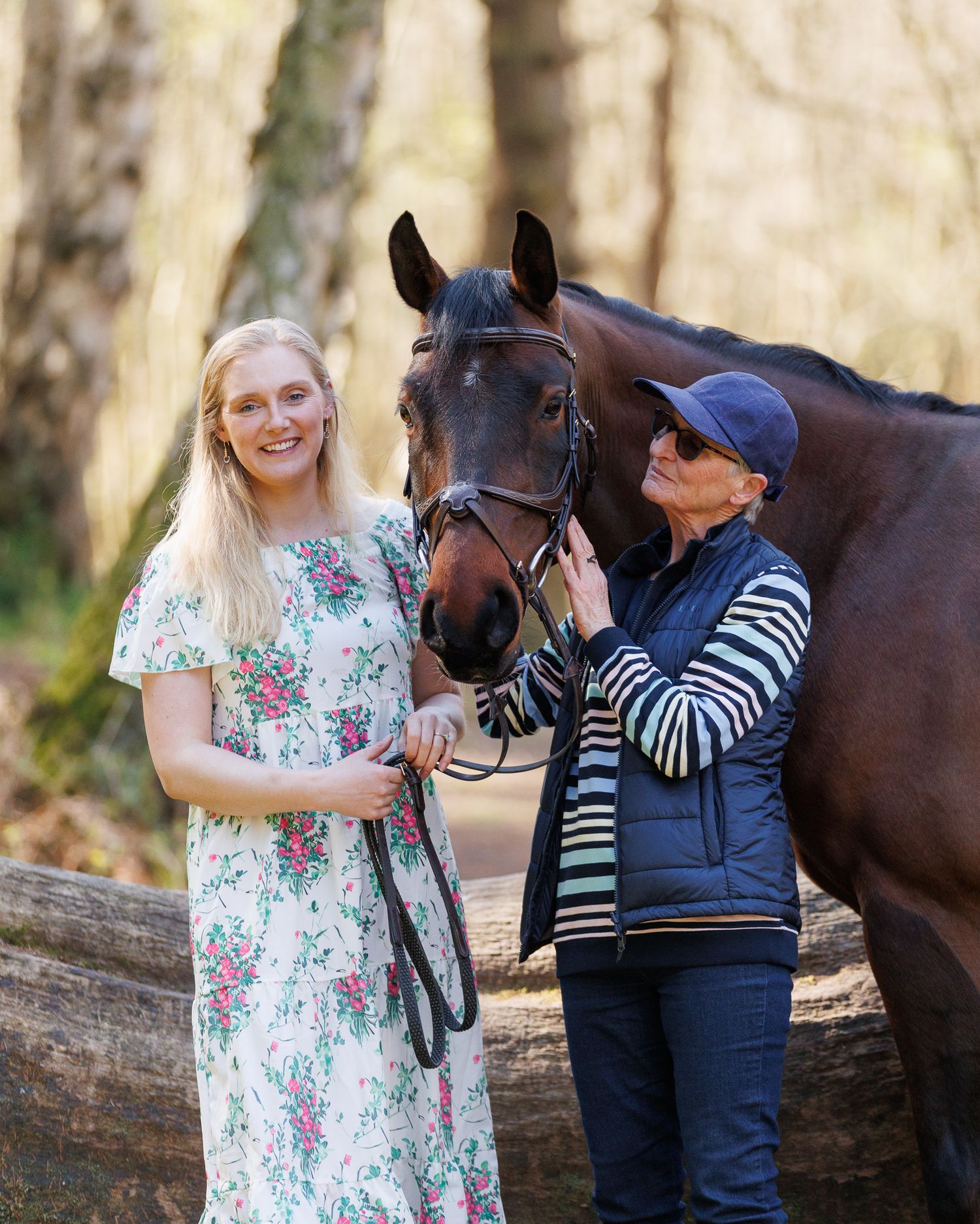 Two women smile beside a dark bay horse in a sunlit forest setting, one in a floral dress, one in a navy vest.