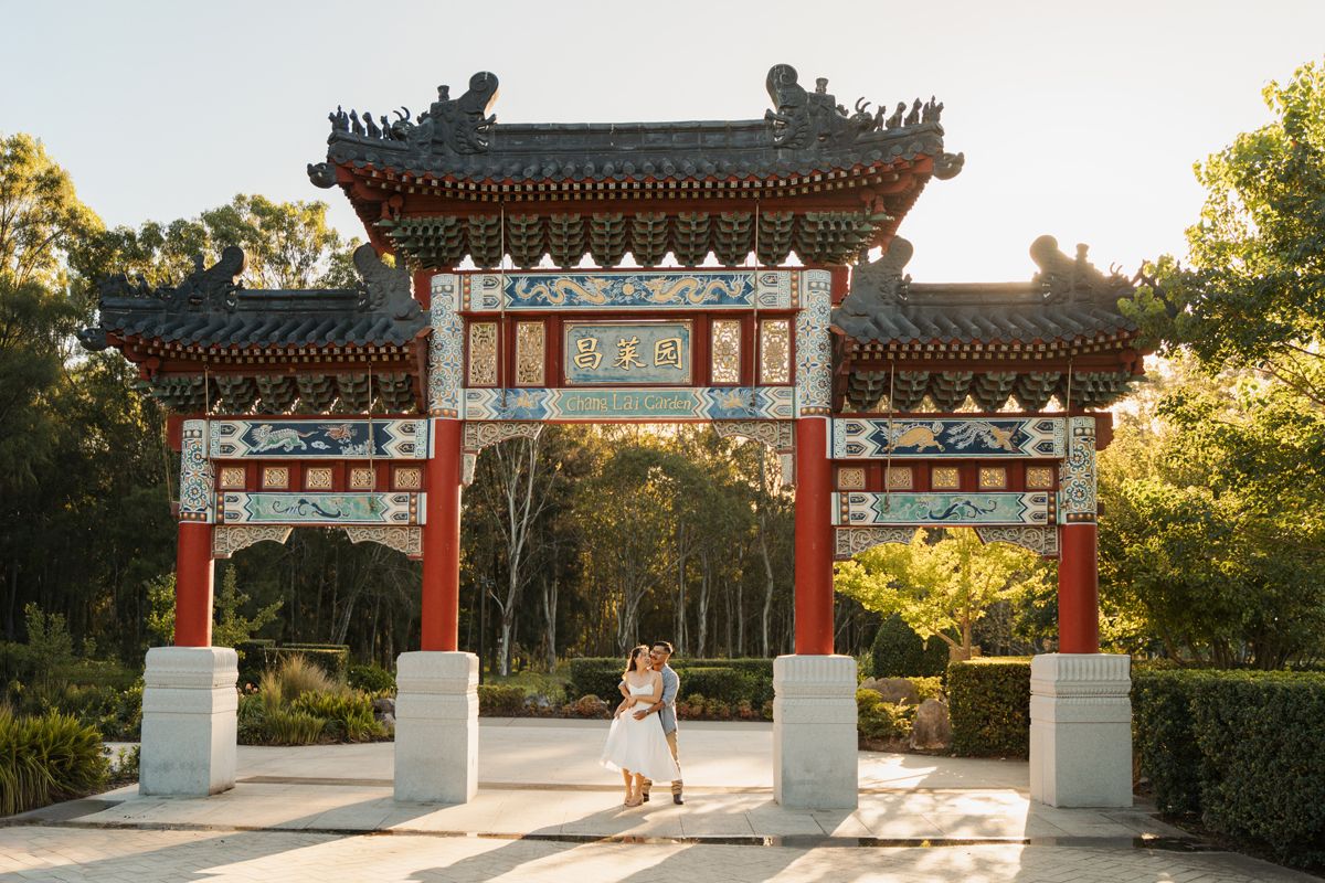 Couple embracing under ornate Chinese paifang gate with red columns and decorative roof in a sunlit Nurragingy reserve photoshoot