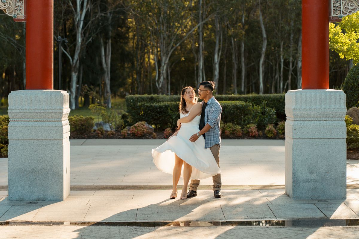 Couple dancing joyfully between red pillars in a sunlit western sydney garden with green trees in the background during couples photoshoot