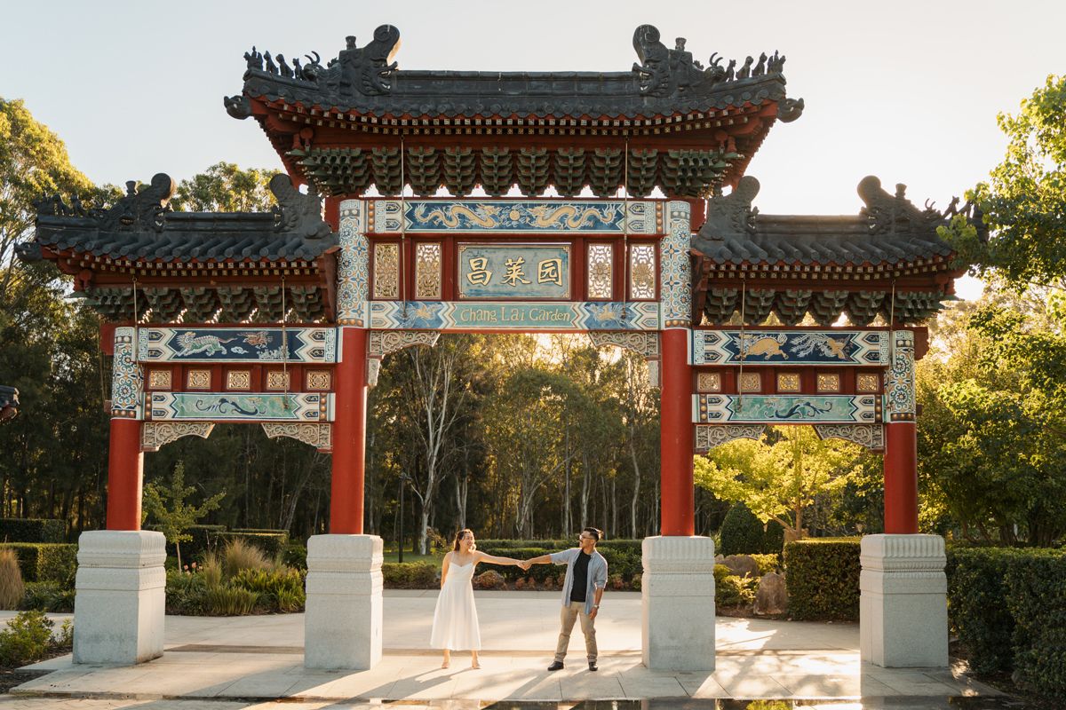 Couple standing beneath ornate red and gold Chinese ceremonial gate surrounded in golden sunlight in western sydney parklands couples photos