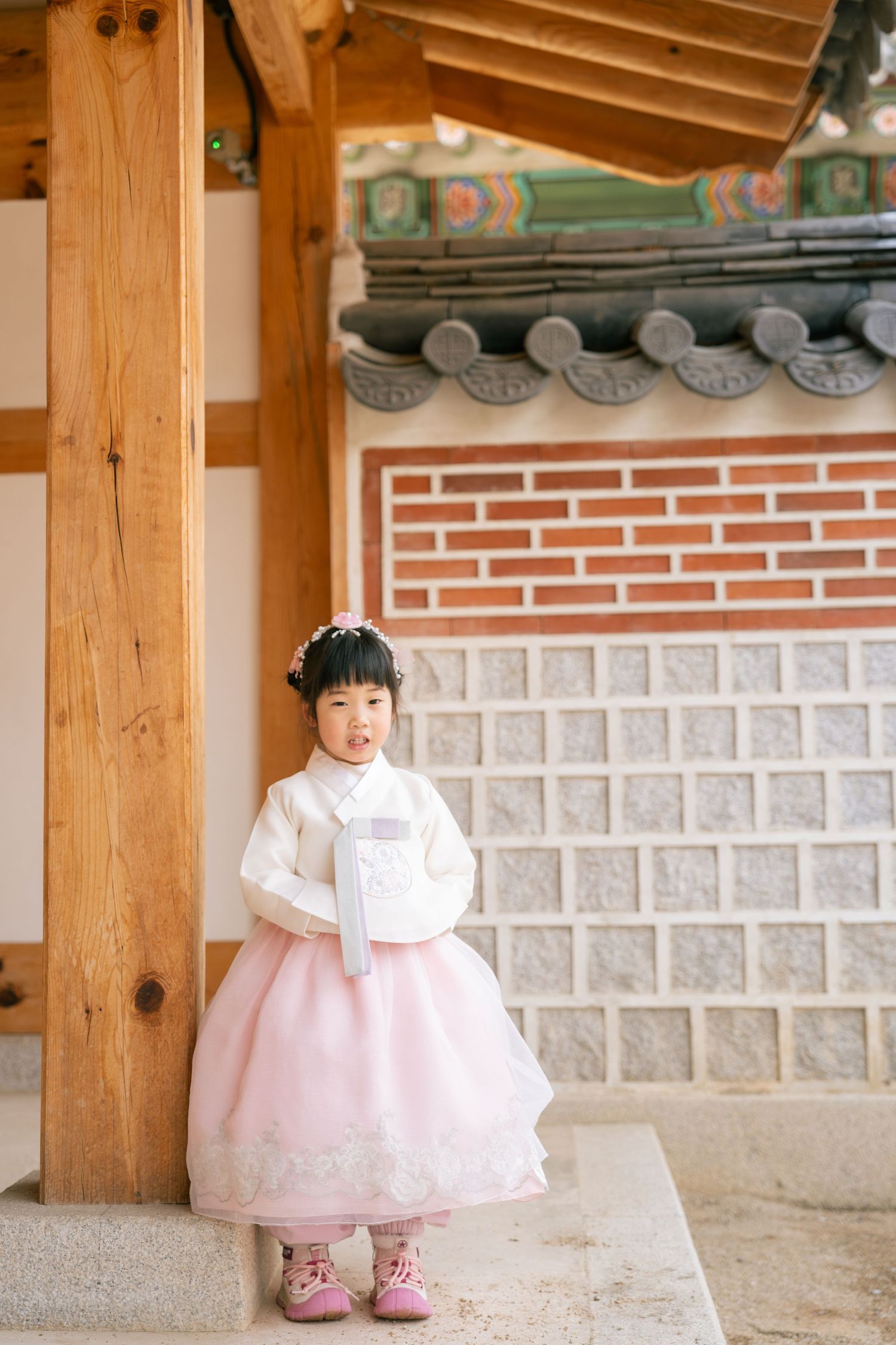 A young child in a traditional Korean hanbok dress with pink skirt stands in front of a temple wall with ornate roof tiles.