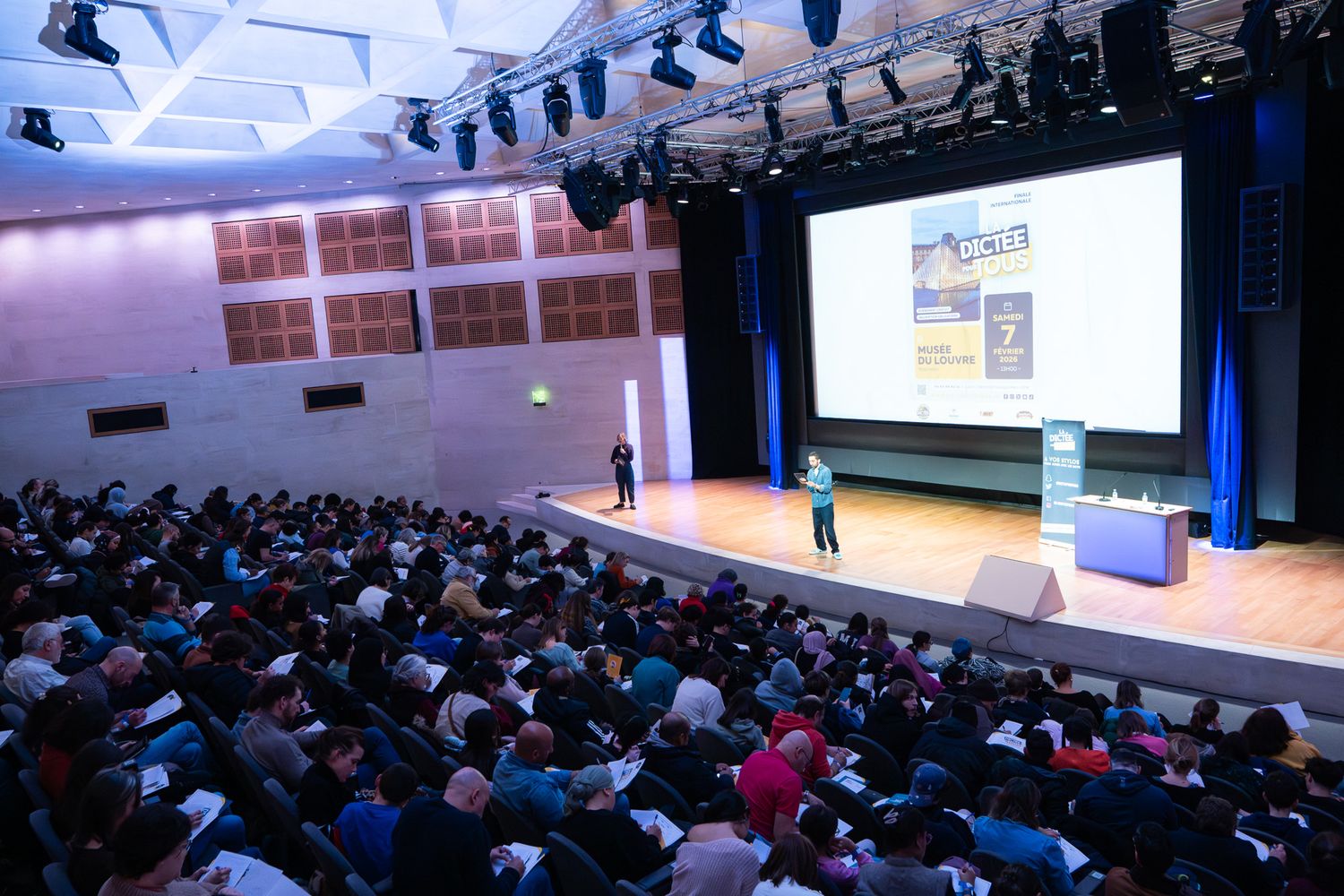 Large audience seated in a conference hall during a business presentation with multiple screens visible on stage.