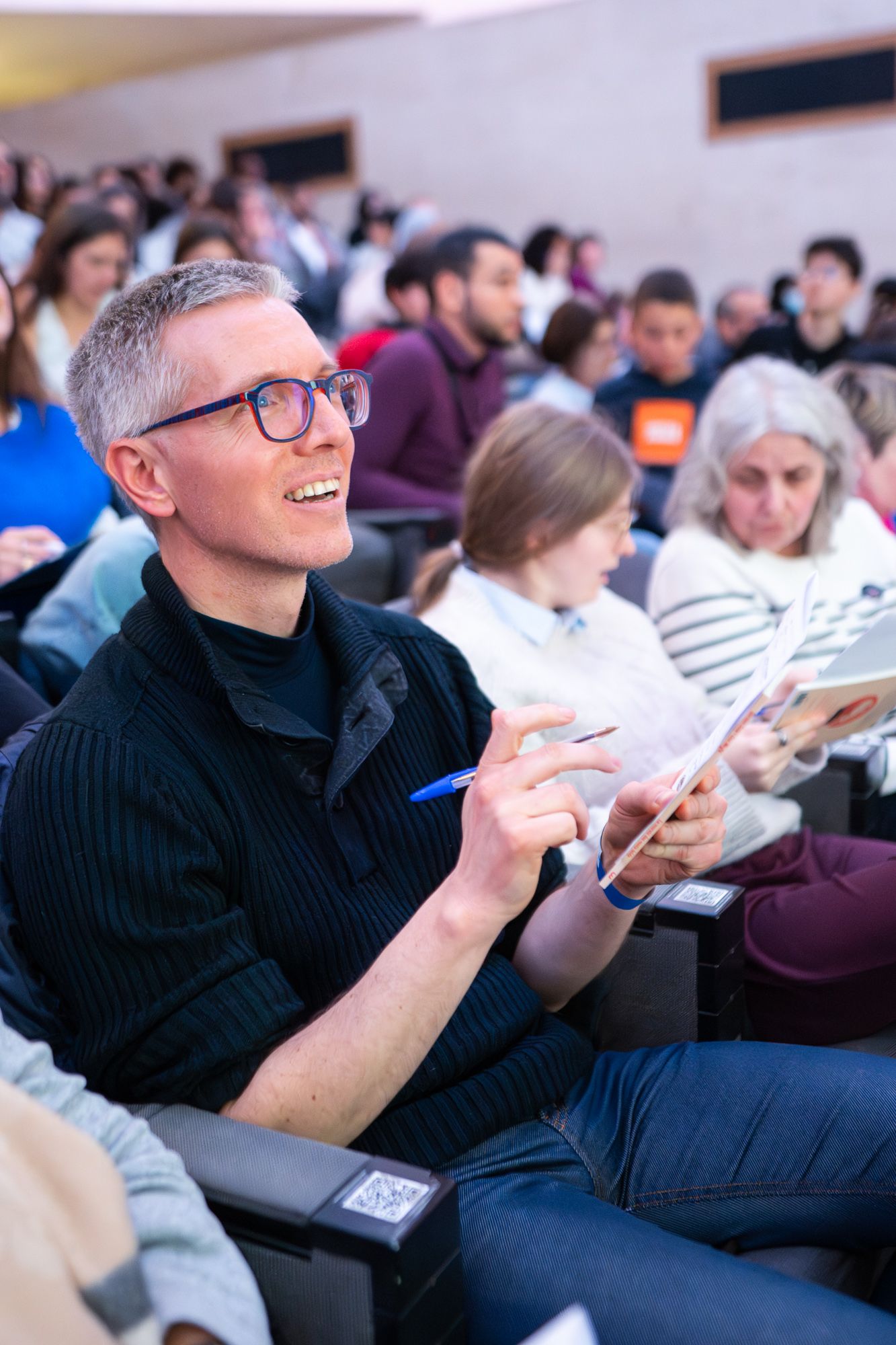A person in a black sweater takes notes while sitting in a crowded lecture hall or conference room.