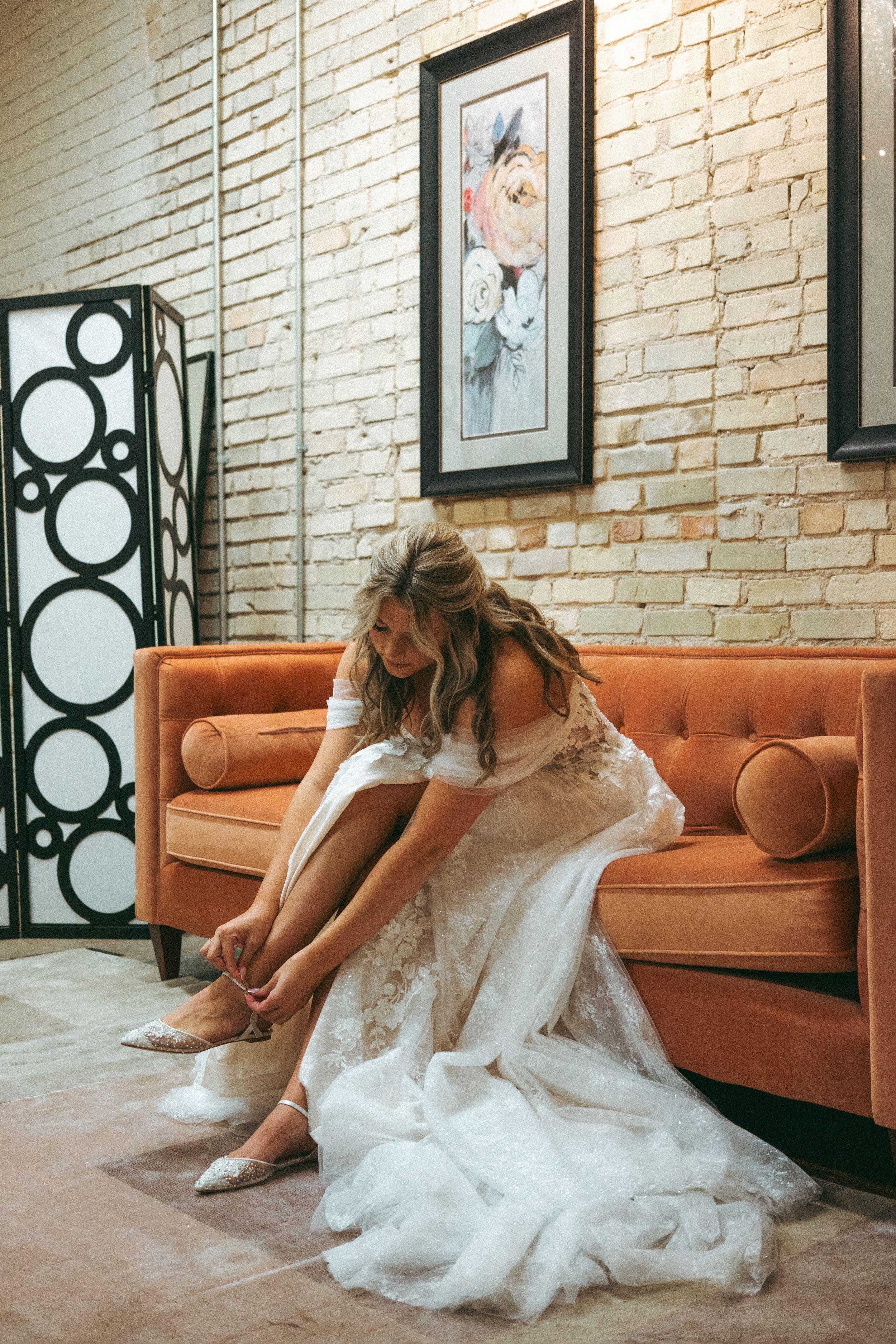 Bride in a lace wedding dress adjusting her heels while seated on an orange velvet sofa indoors.