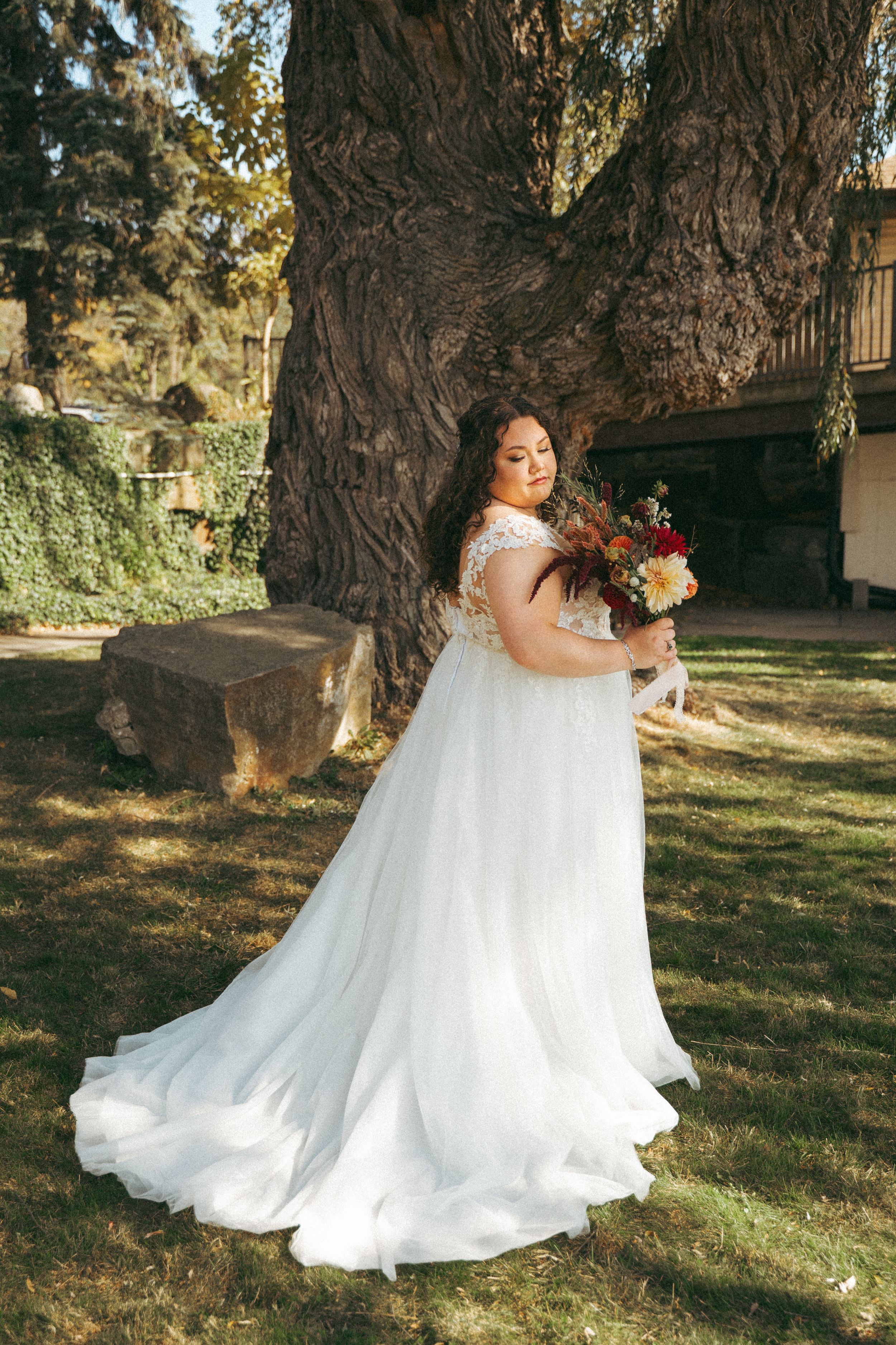 Plus-size bride in a flowing white lace wedding gown holding a bouquet beside a large tree outdoors.