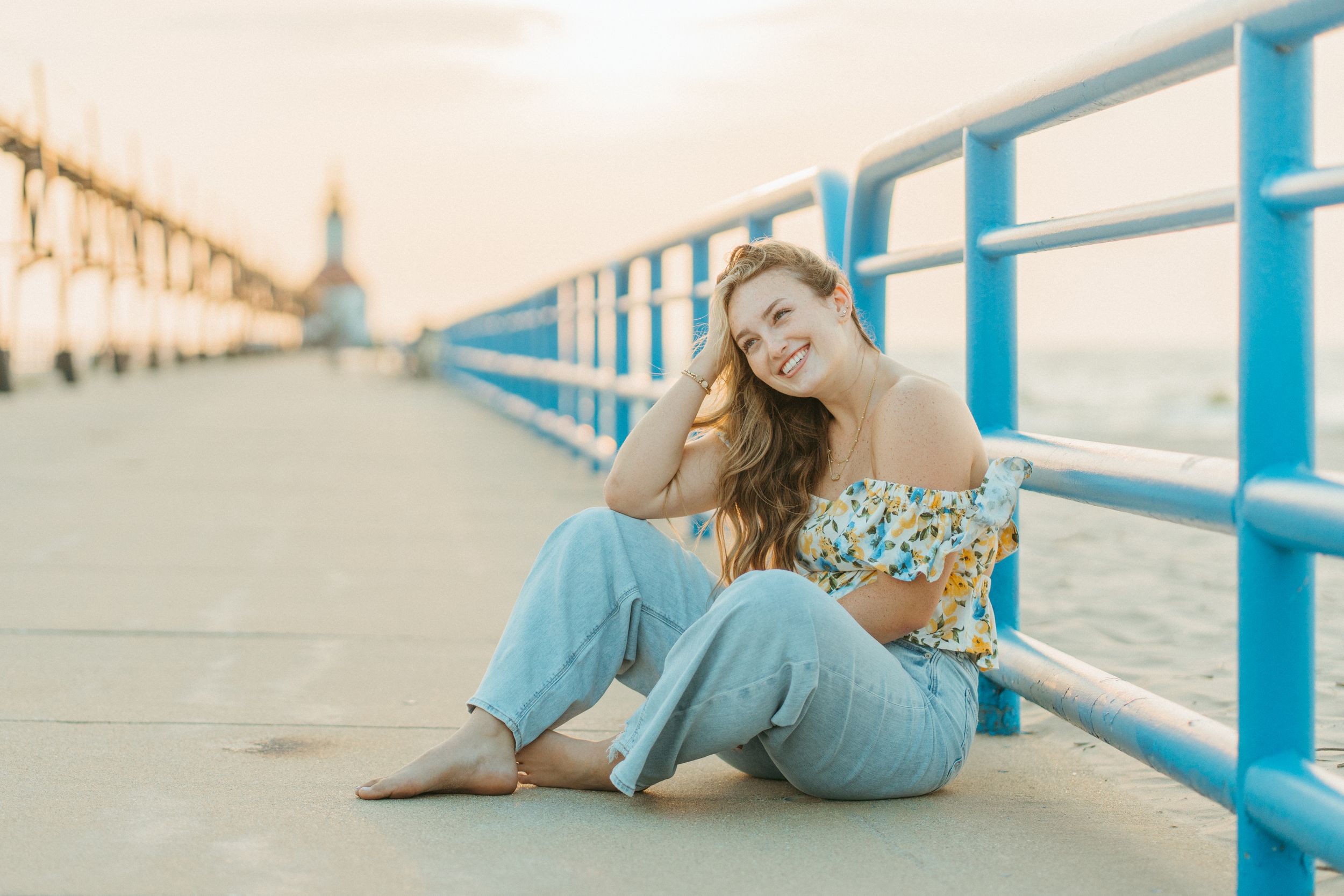 Smiling young woman sitting on a pier boardwalk at sunset with blue railings and a lighthouse behind her.