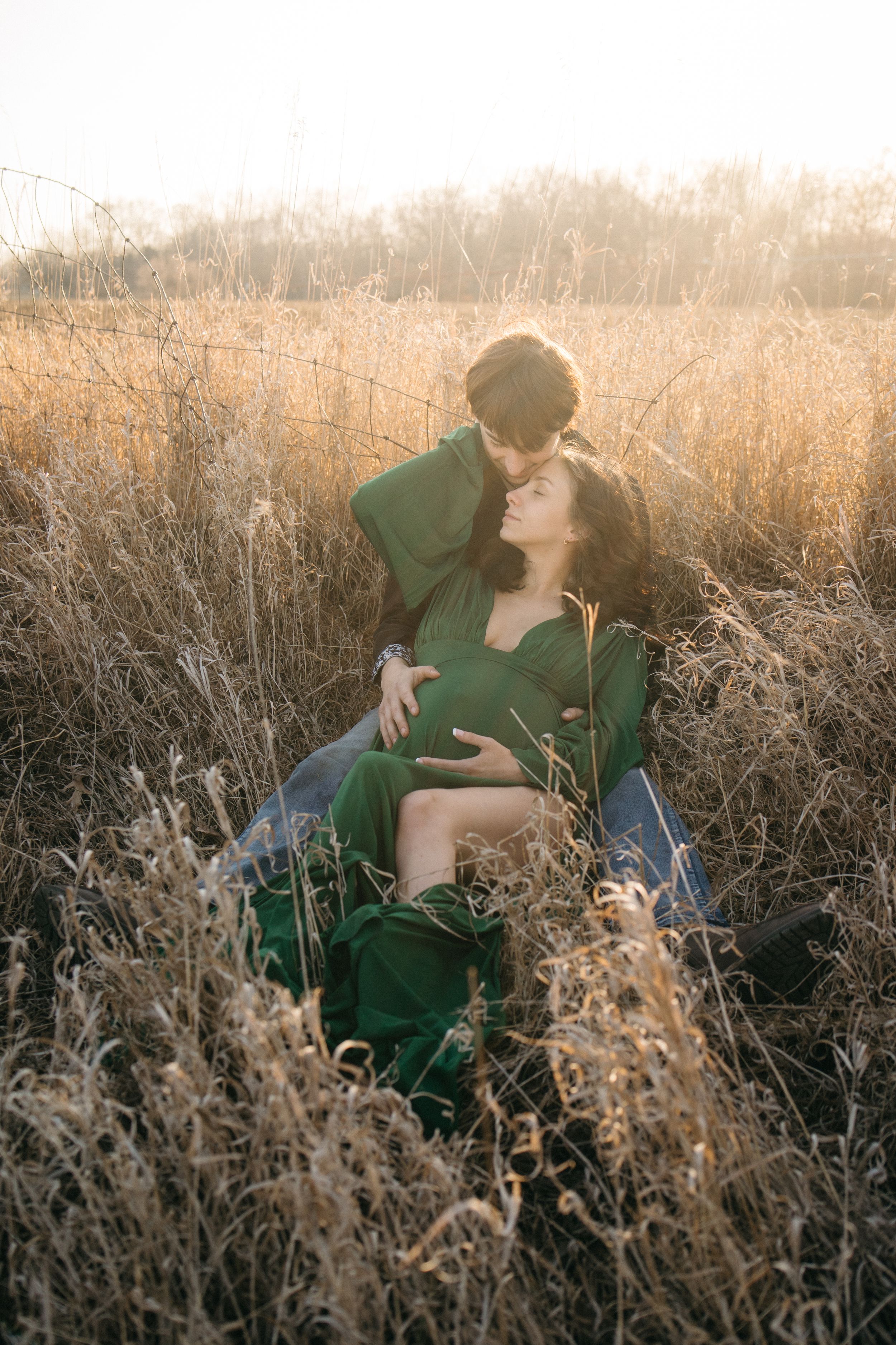 Pregnant woman in green dress reclines against partner in golden wheat field at sunset.