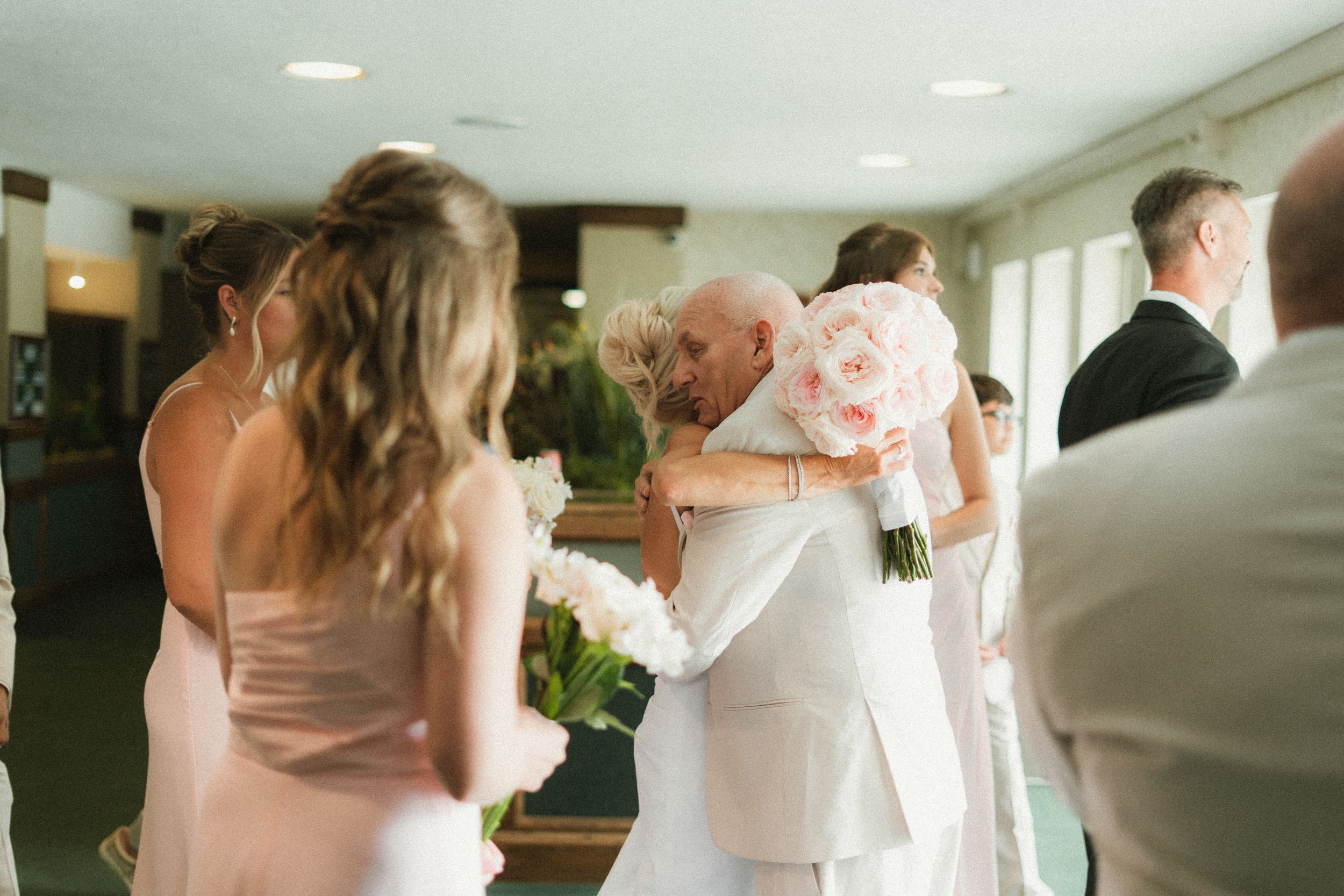 Bride hugging elderly guest in white suit at indoor wedding reception while bridesmaids hold floral bouquets.