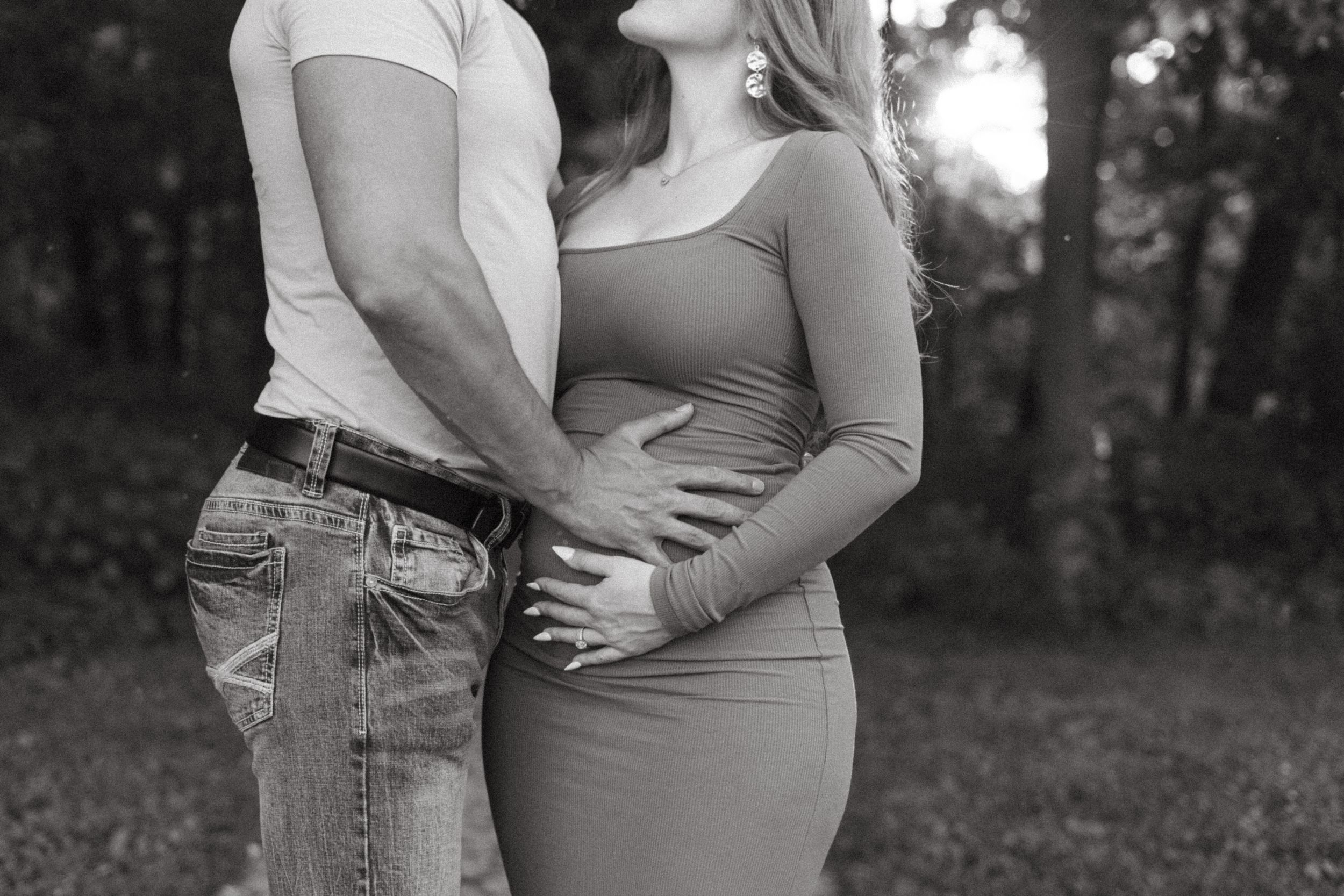 Black and white maternity photo of couple embracing outdoors, hands cradling pregnant belly.